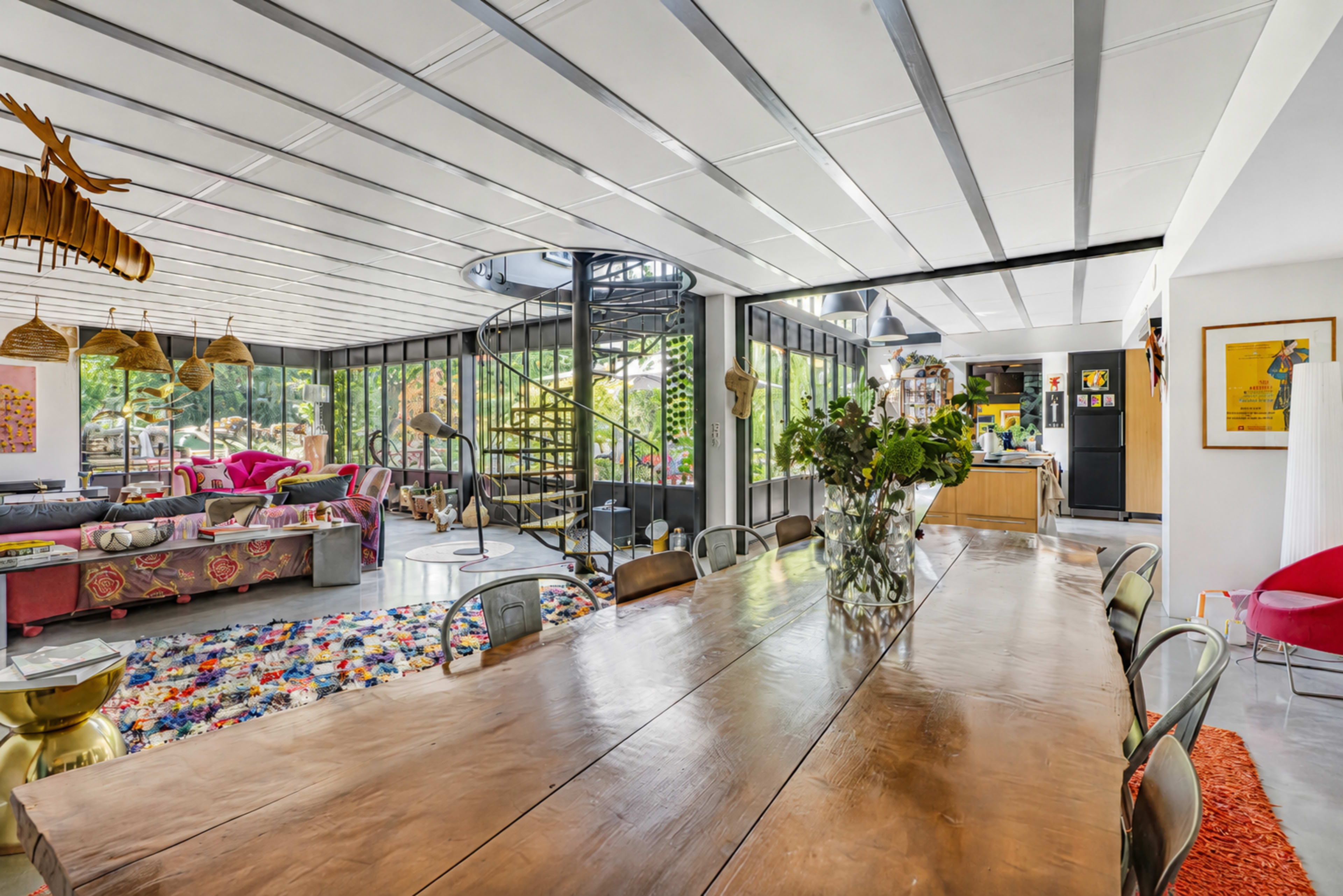 The image shows a spacious, modern dining area with a large wooden table, surrounded by metal chairs, and featuring large glass windows that open to a greenery-filled patio.