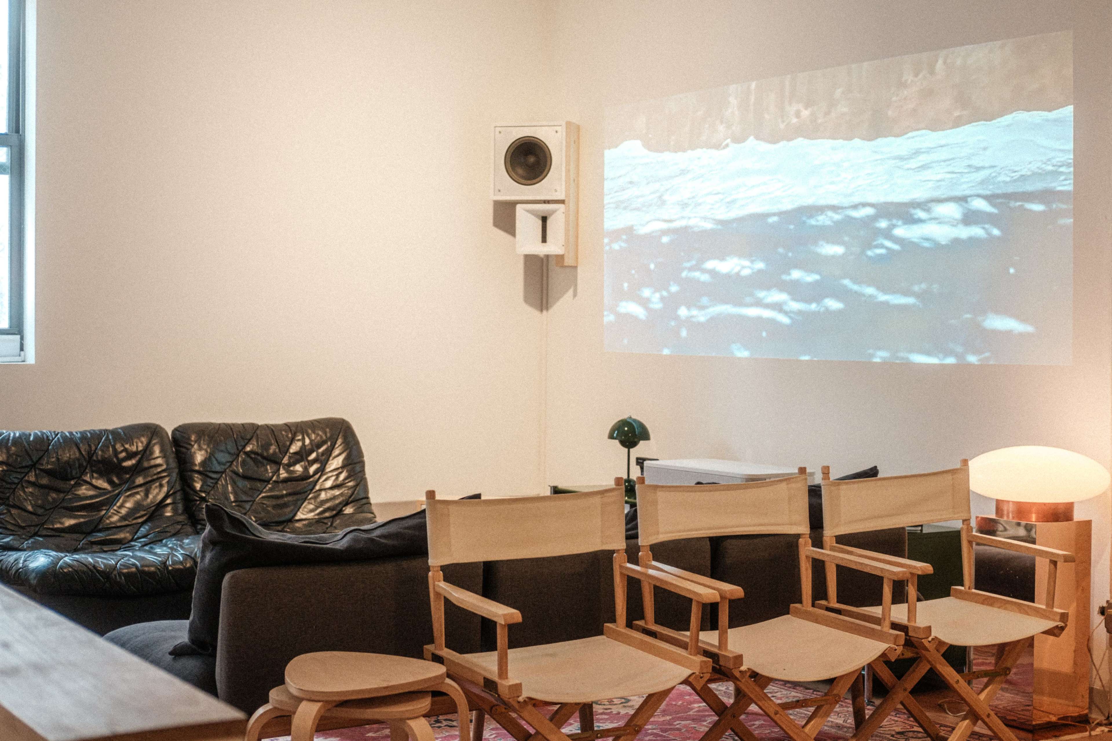 The image shows a cozy living room with modern furniture, including two couches, a small table, and director's chairs, along with a projection of water on the wall.