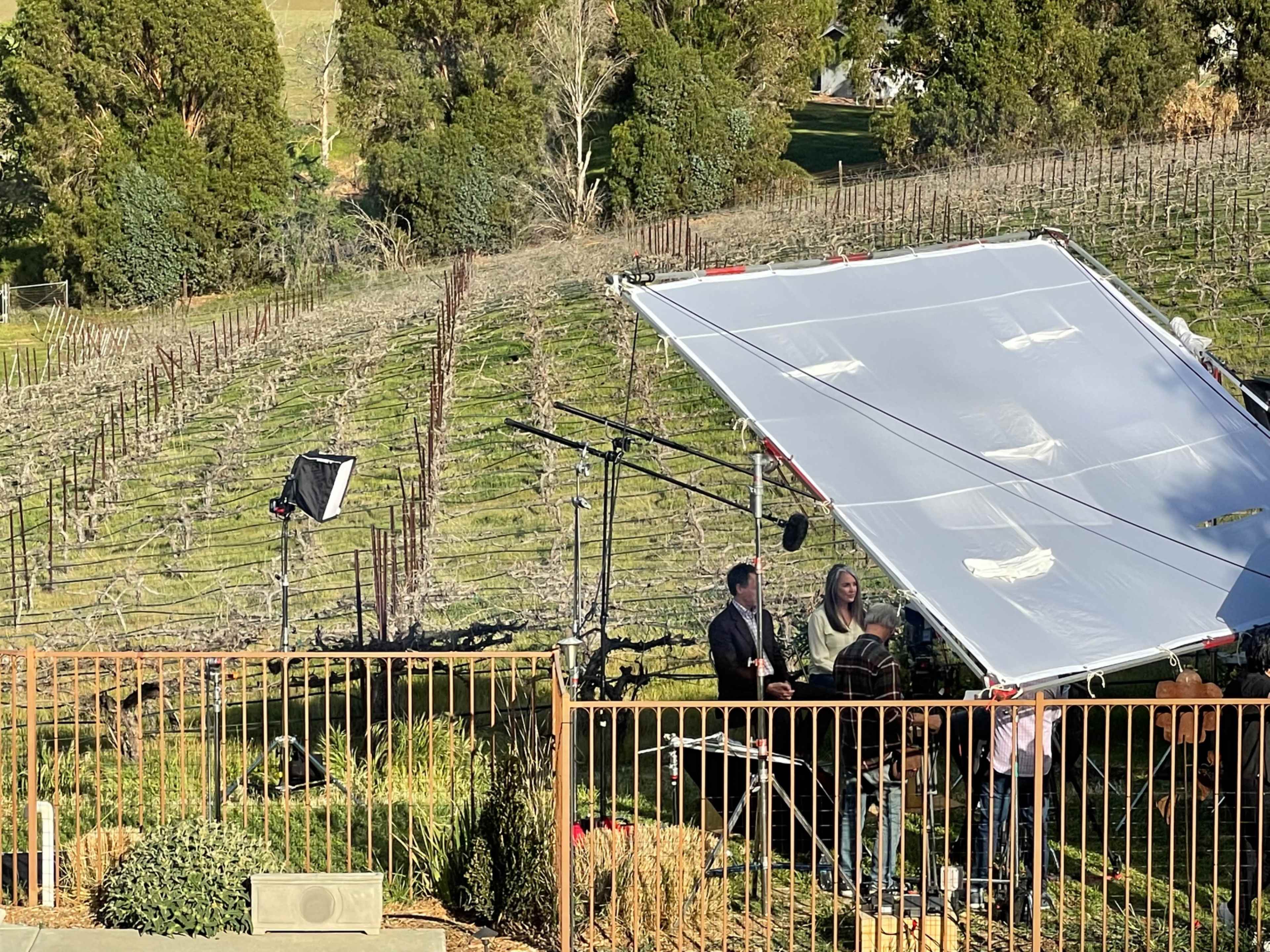 A film crew sets up a shoot in a vineyard with large equipment positioned under a shaded canopy.
