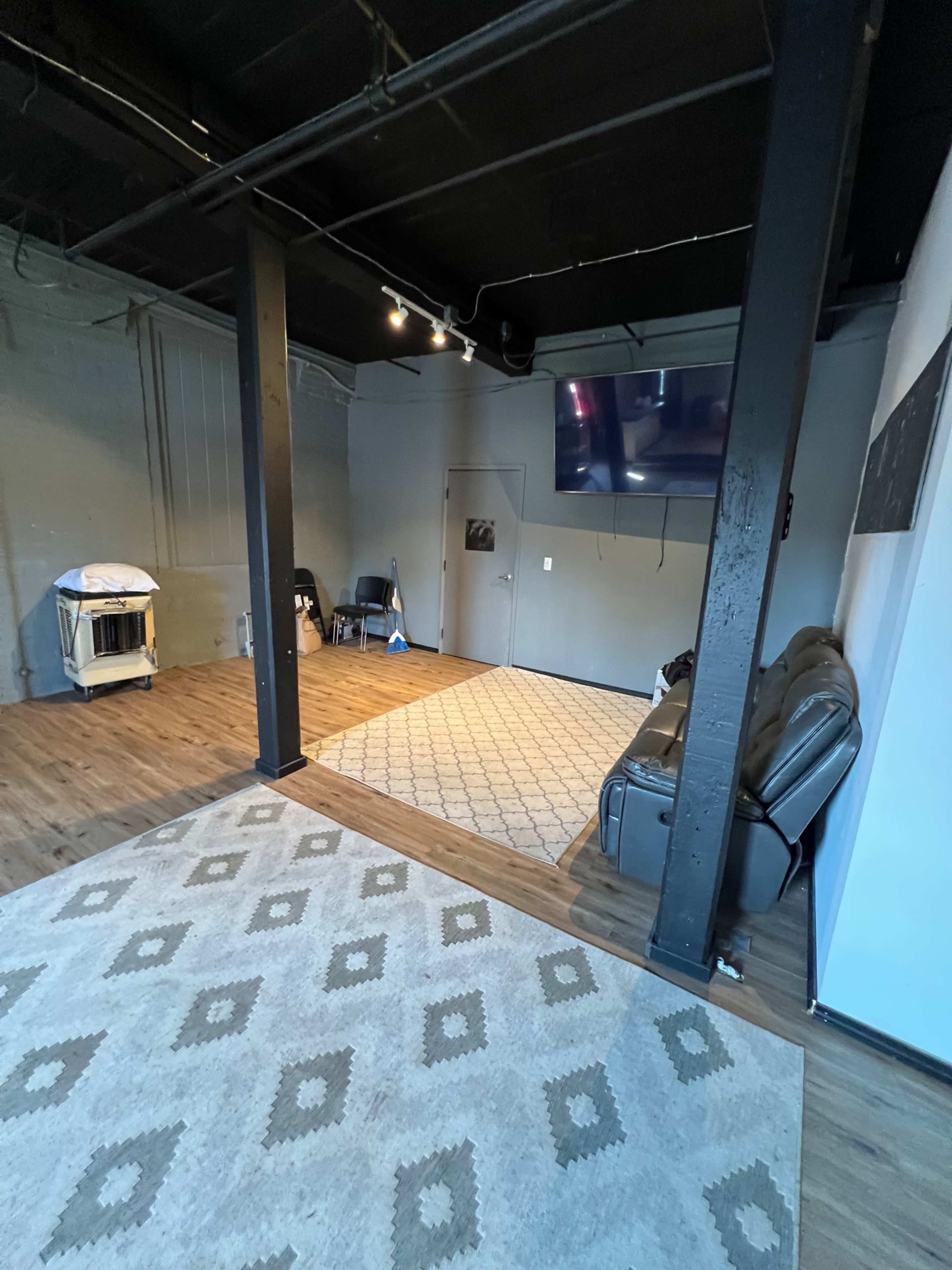 A sparsely furnished room with a gray wall, wooden flooring, a large area rug, a leather couch, and a wall-mounted television.
