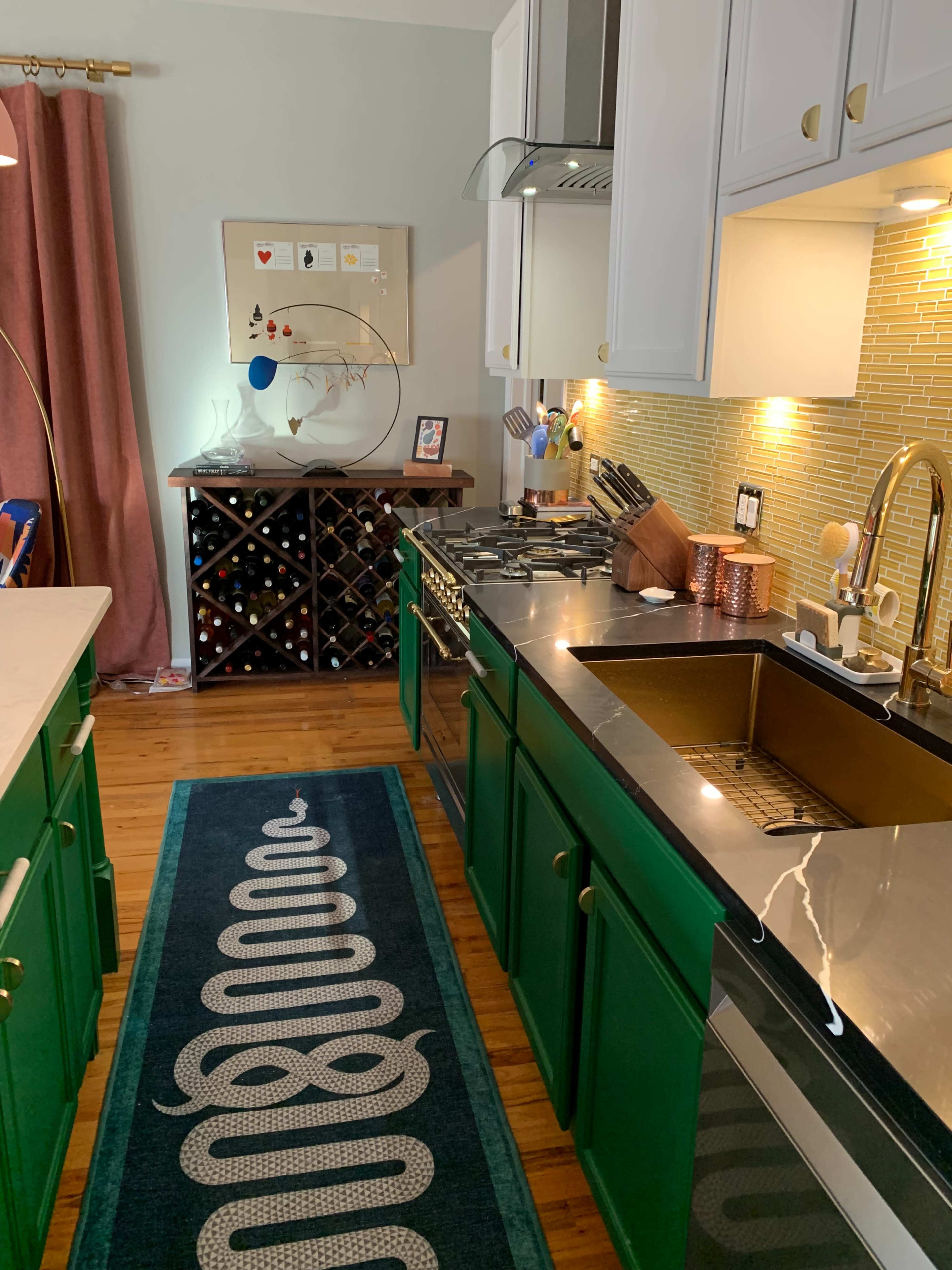 A modern kitchen features green cabinetry, a stainless steel countertop, and a backsplash of yellow tiles, with a dark wooden wine rack and a rug laid along the floor.