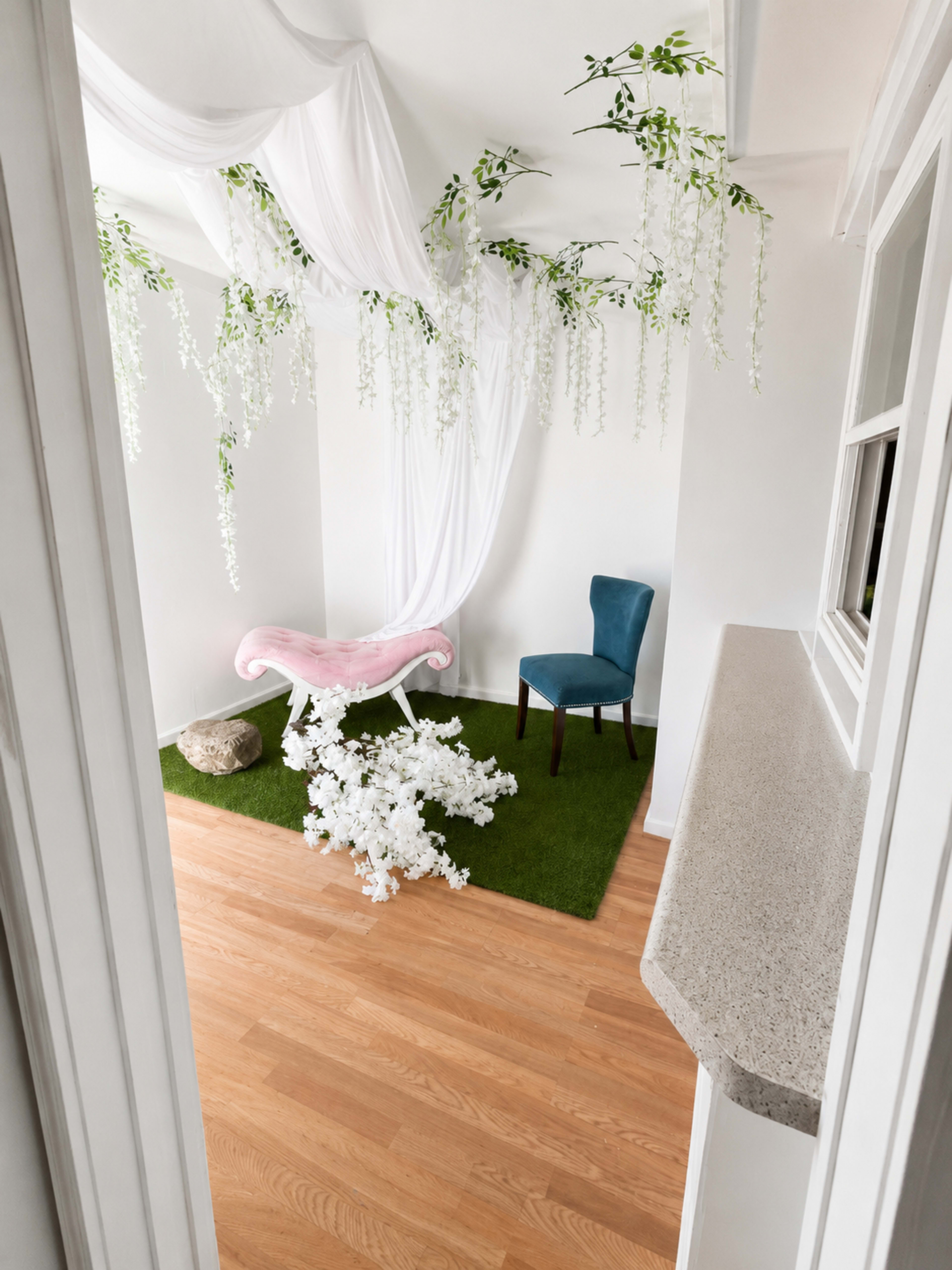 The room features a pink chaise lounge under draped white fabric, surrounded by green artificial grass and white flower arrangements.