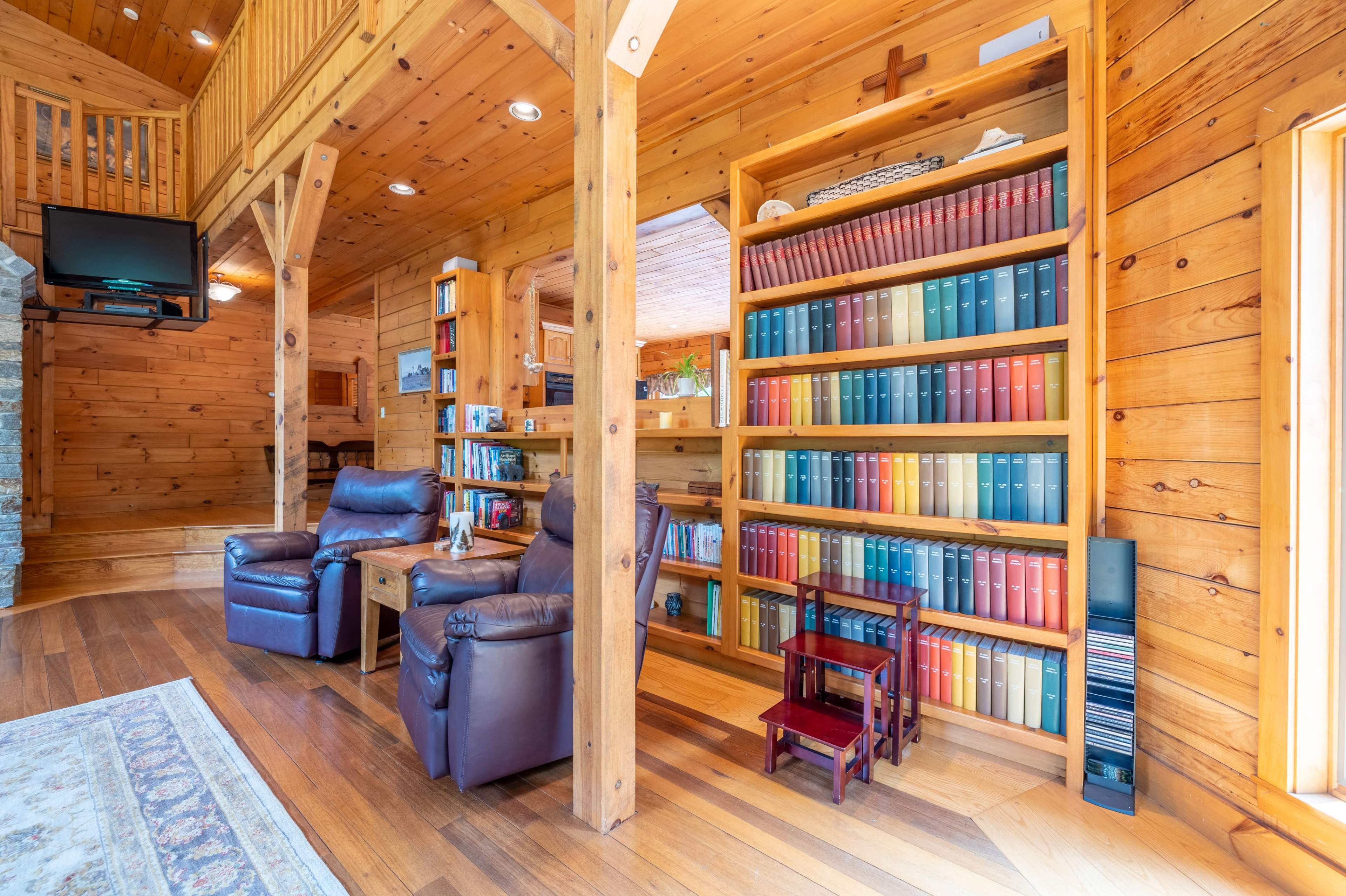 A wooden interior of a cozy room featuring two leather armchairs, a small table, and a tall bookshelf filled with colorful books.