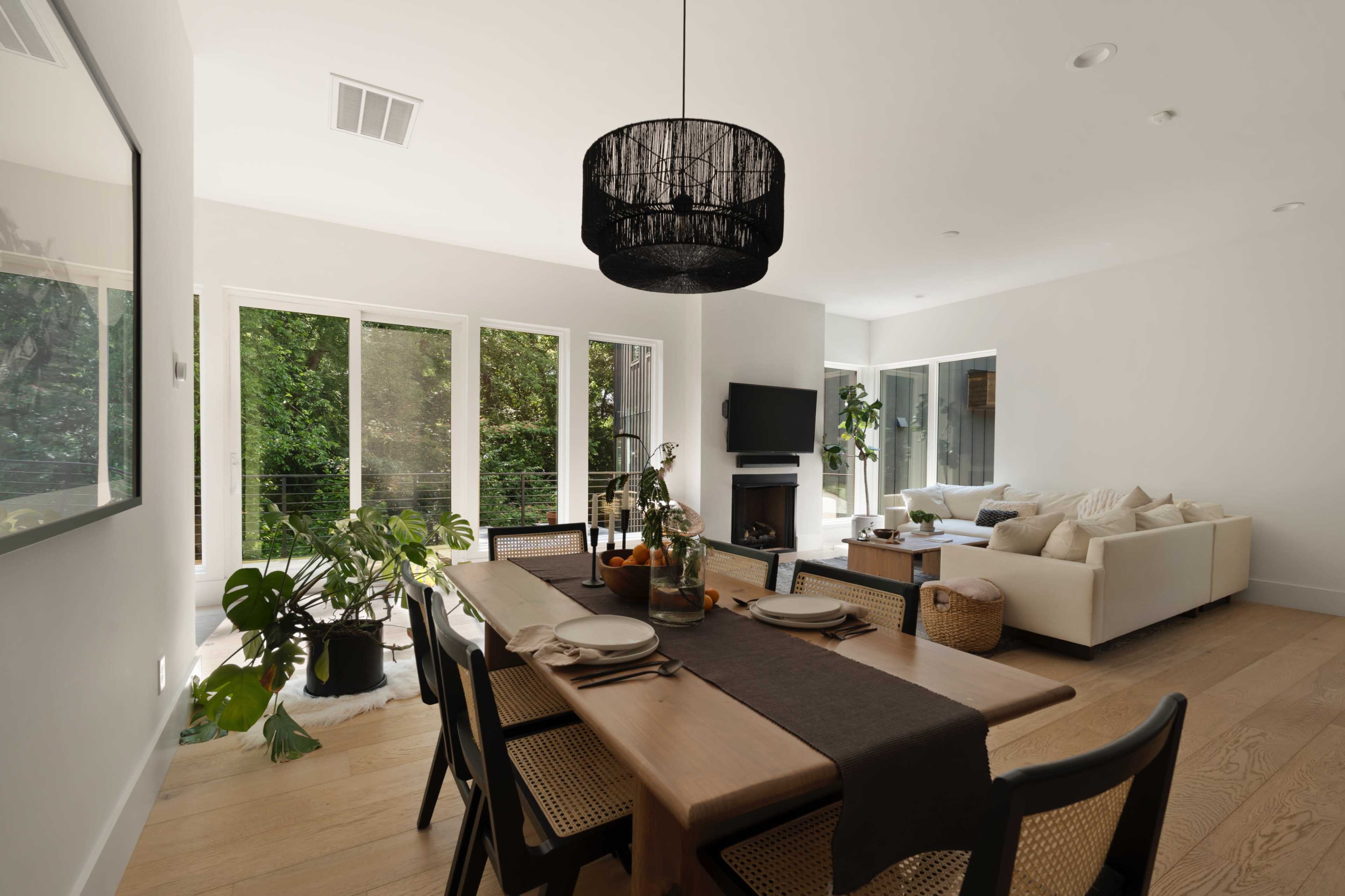 A dining area featuring a wooden table, black chairs, and a chandelier, with large windows overlooking greenery and a living room visible in the background.