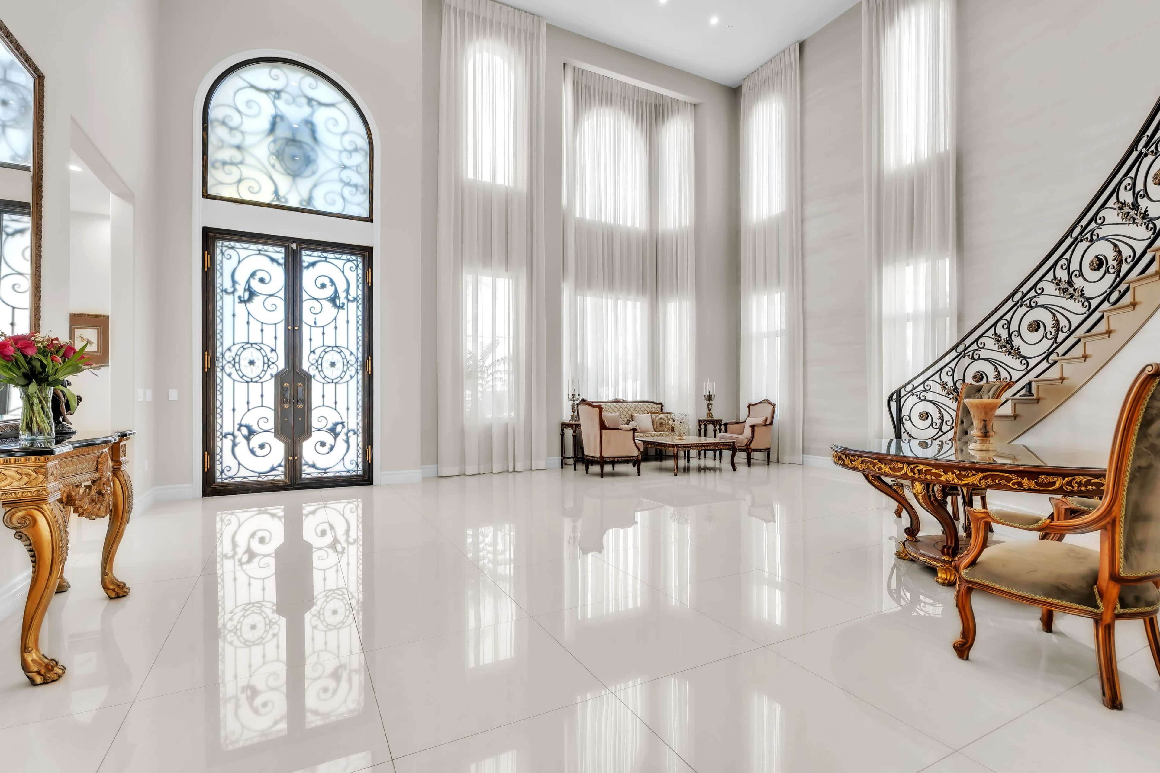 The image depicts a spacious foyer with high ceilings, large windows, and elegant furnishings, including a staircase and a decorative table.