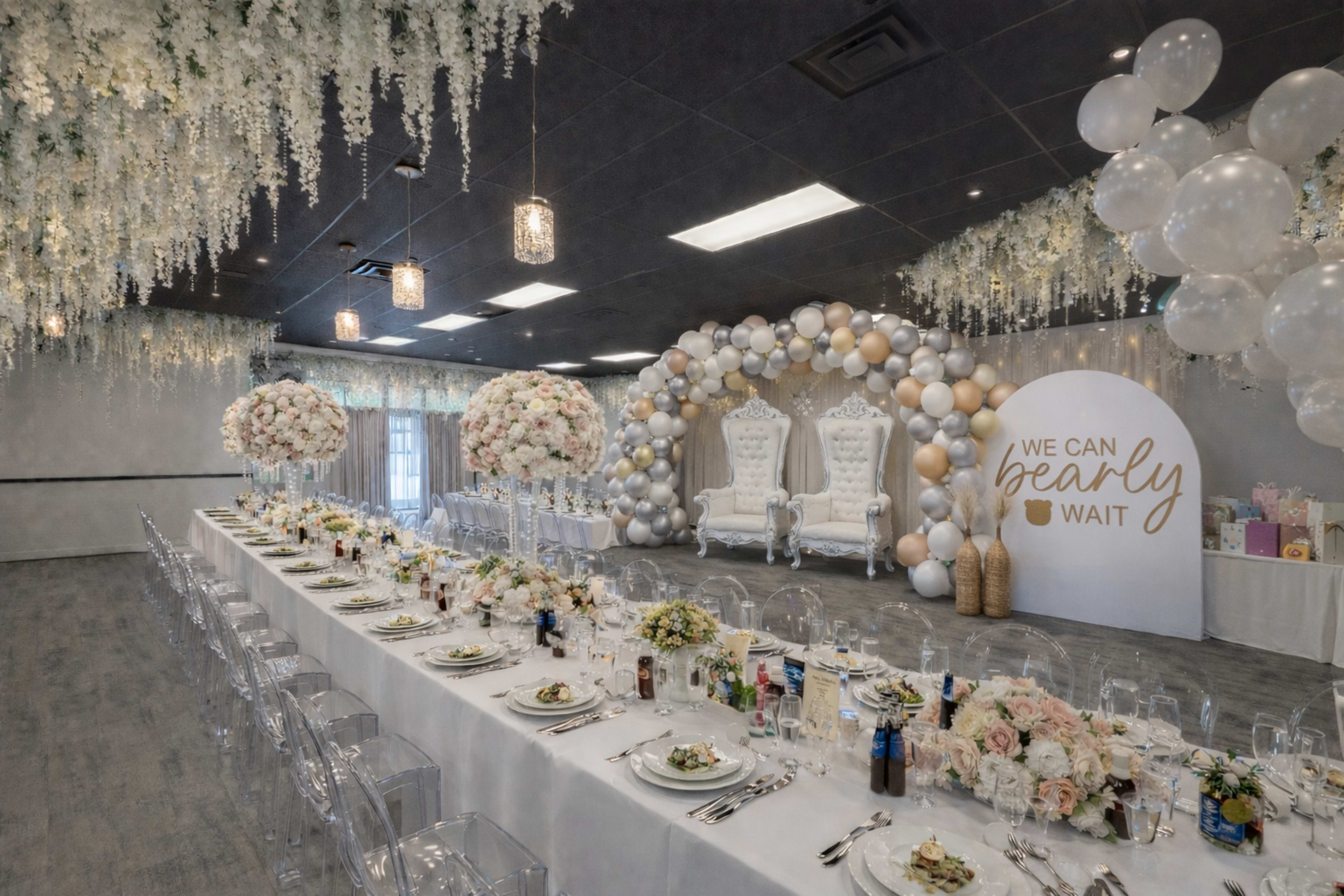 A beautifully arranged banquet hall features a long dining table set with elegant tableware, surrounded by floral centerpieces and decorated with balloons, while a backdrop displays two ornate chairs and a sign reading "WE CAN BEARLY WAIT."