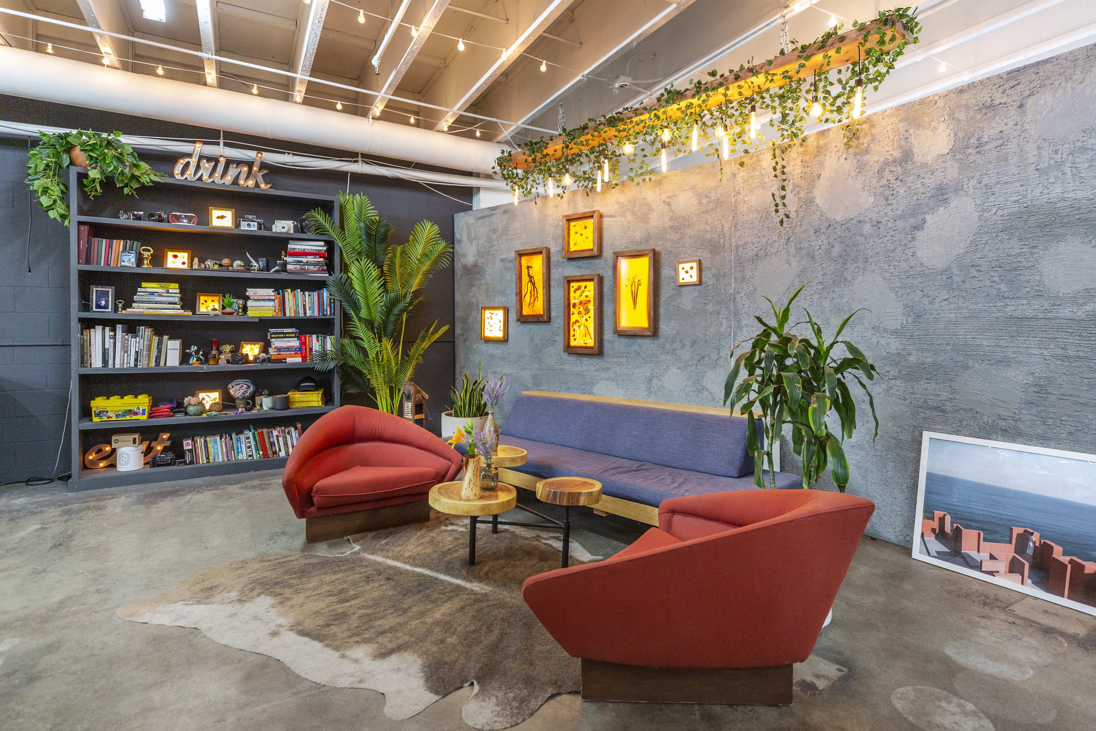 A modern lounge area with two red lounge chairs and a blue sofa arranged around a wooden coffee table, surrounded by plants and shelves filled with books and decorative items.