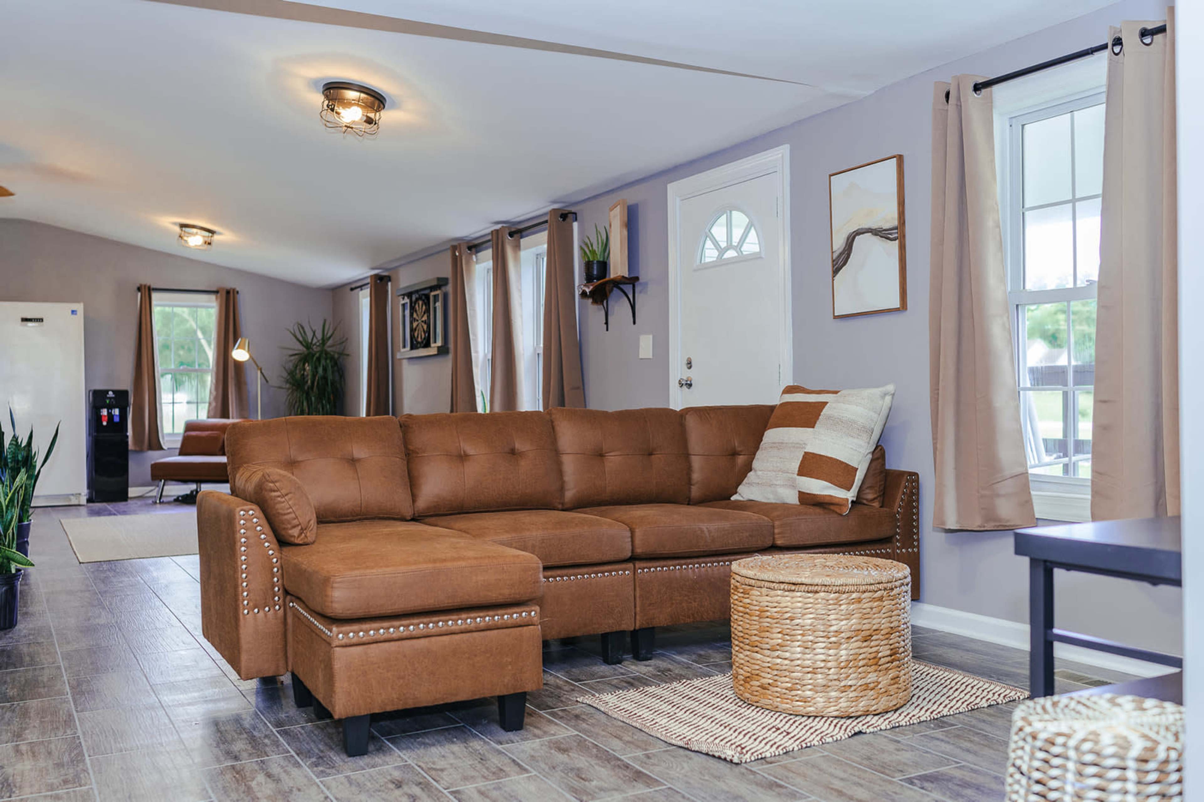 A living room featuring a brown sectional sofa, a round wicker coffee table, and windows dressed with light curtains.