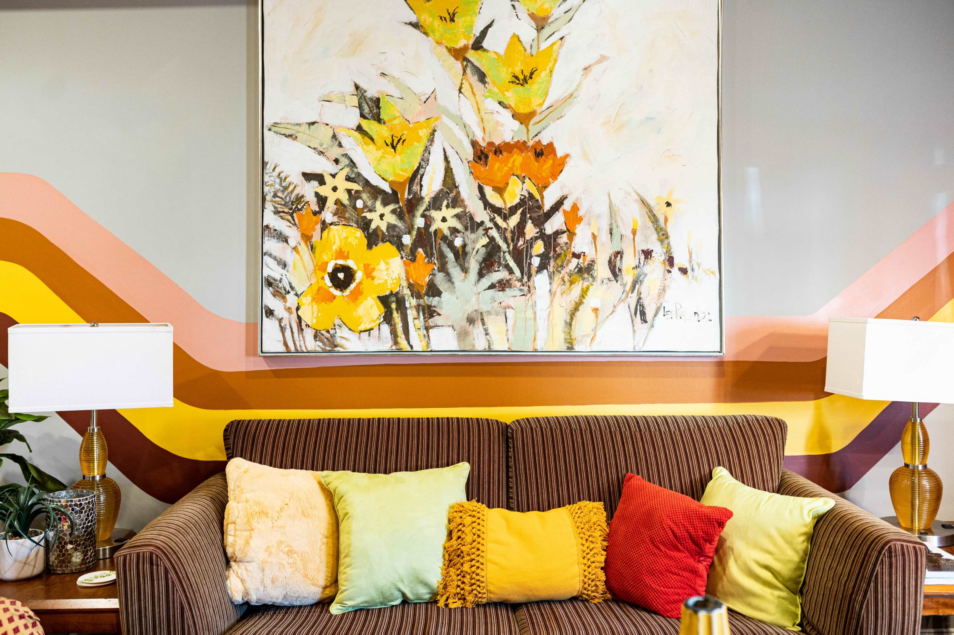 A brown striped sofa adorned with colorful pillows in front of a large floral painting and a wall featuring geometric color blocks.