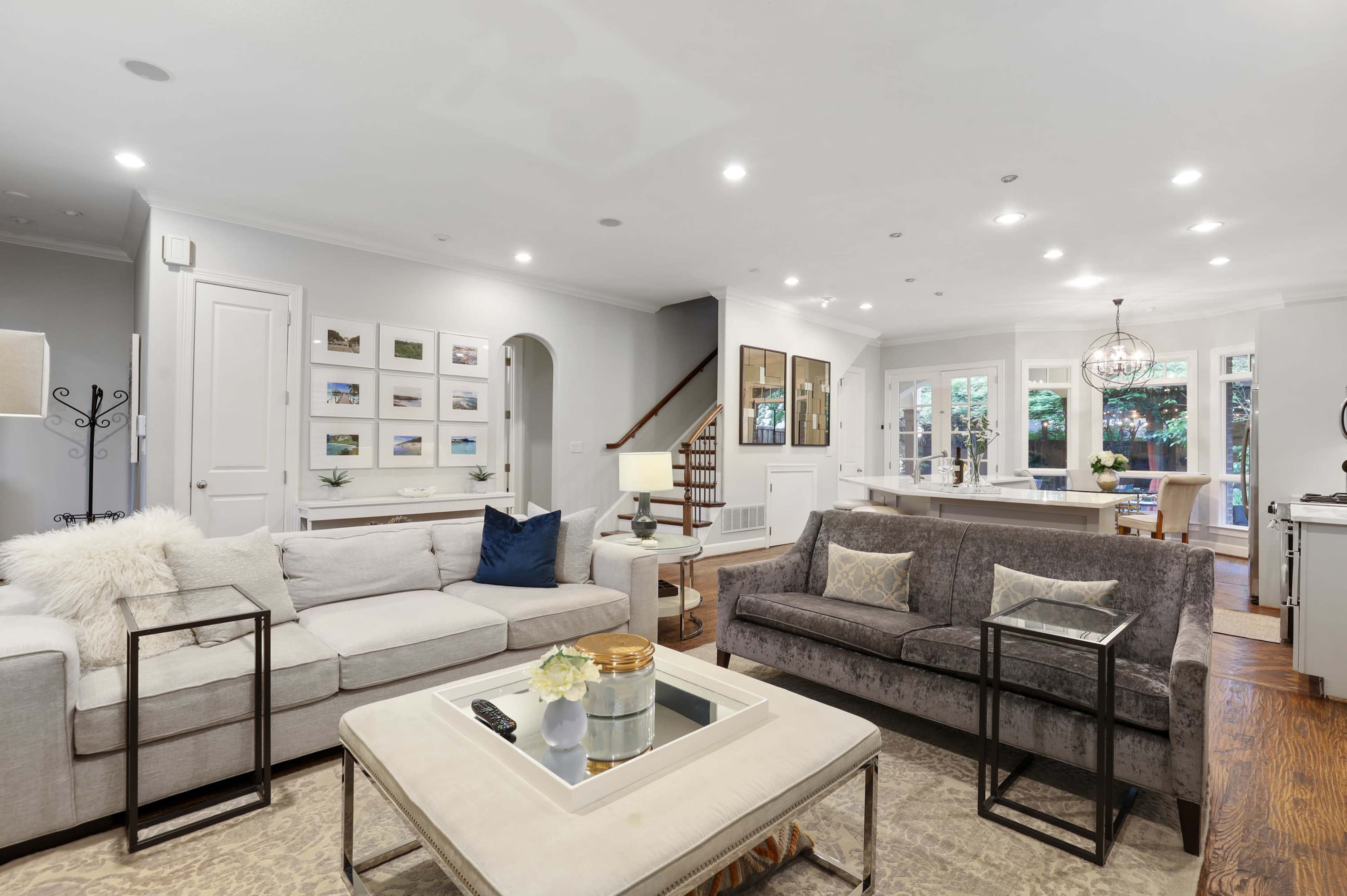 The image shows a spacious living room with two sofas, a coffee table in the center, and a staircase leading to an upper level, all illuminated by recessed lighting.