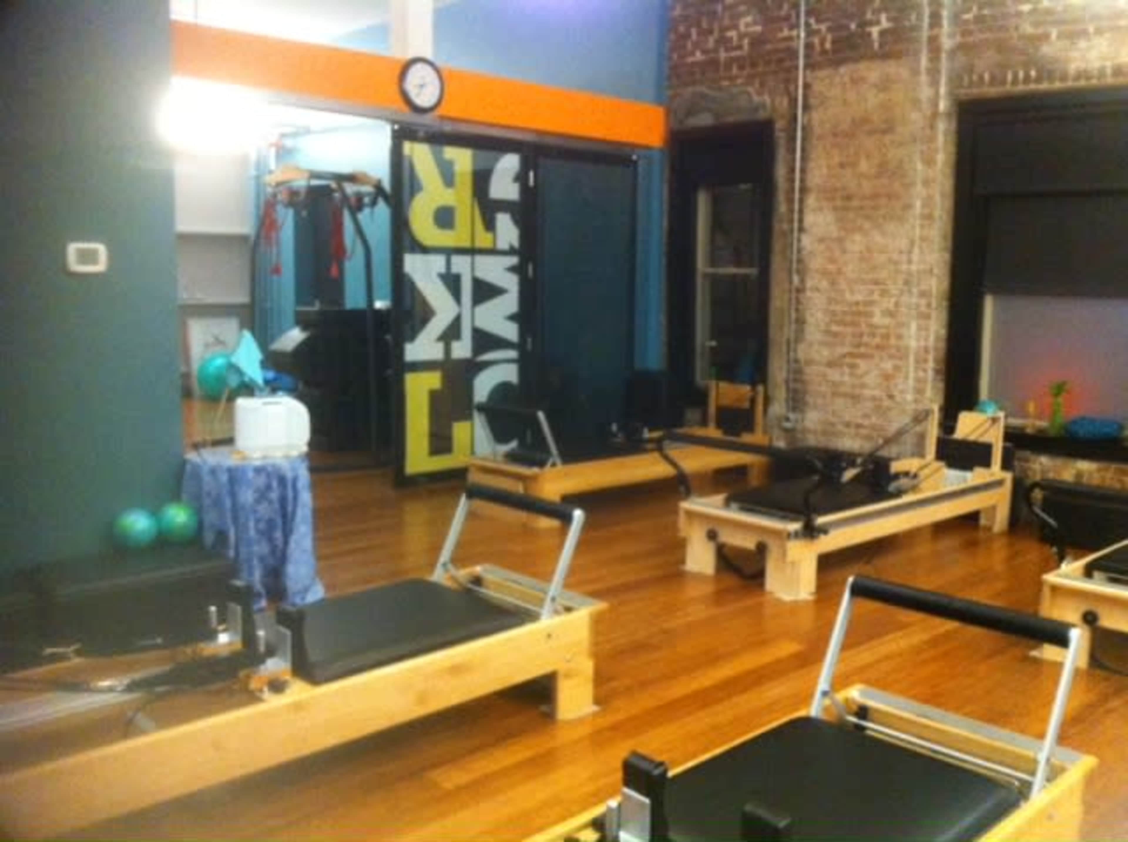A fitness studio with wooden reformer machines arranged on a wooden floor and workout equipment visible in the background.