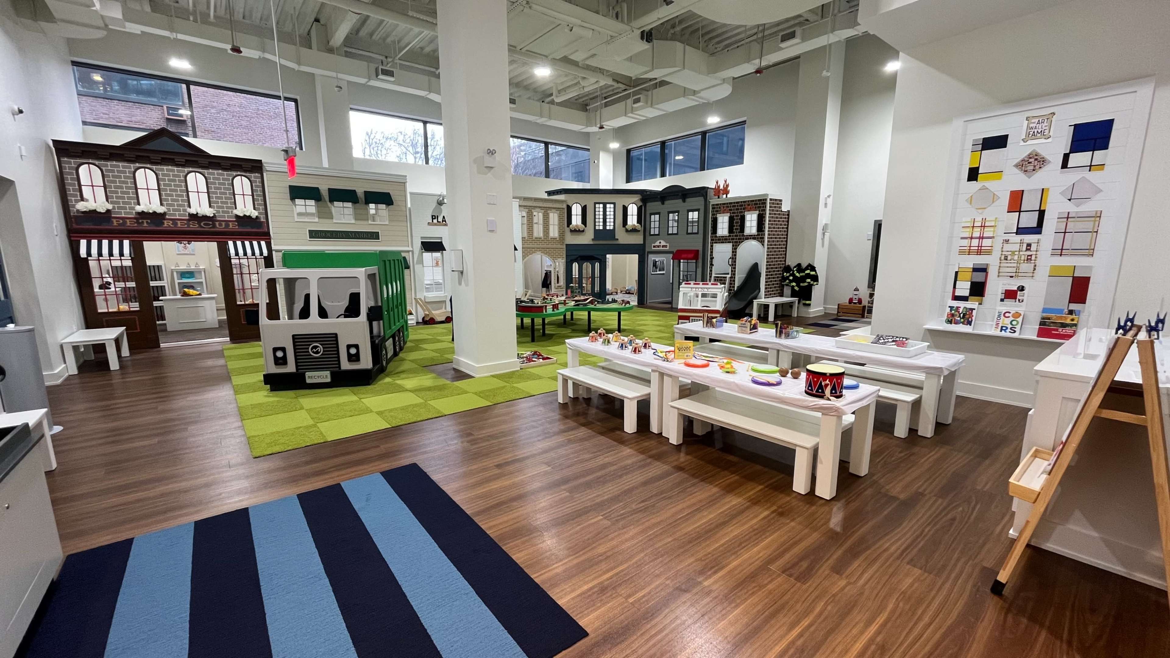 A brightly lit indoor play area designed for children, featuring various play structures, including a bus and storefronts, along with tables for activities.