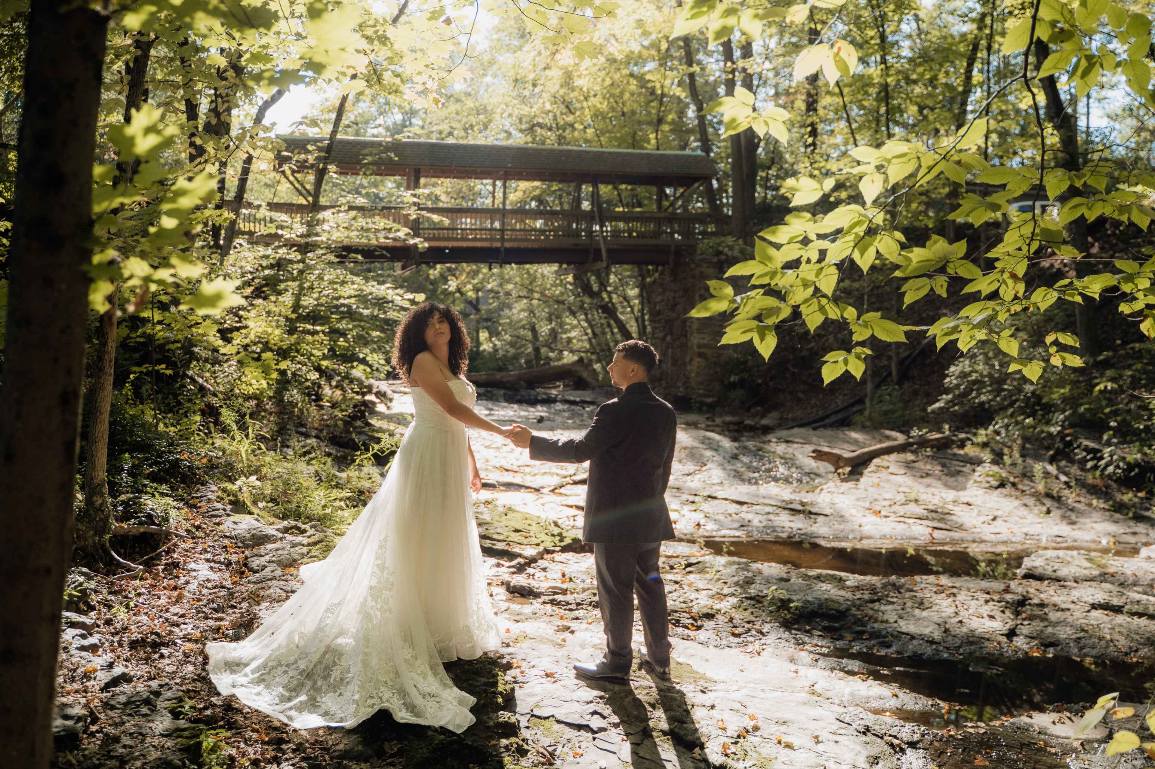 A couple stands hand-in-hand in a sunlit forest by a stream, with a wooden bridge visible in the background.