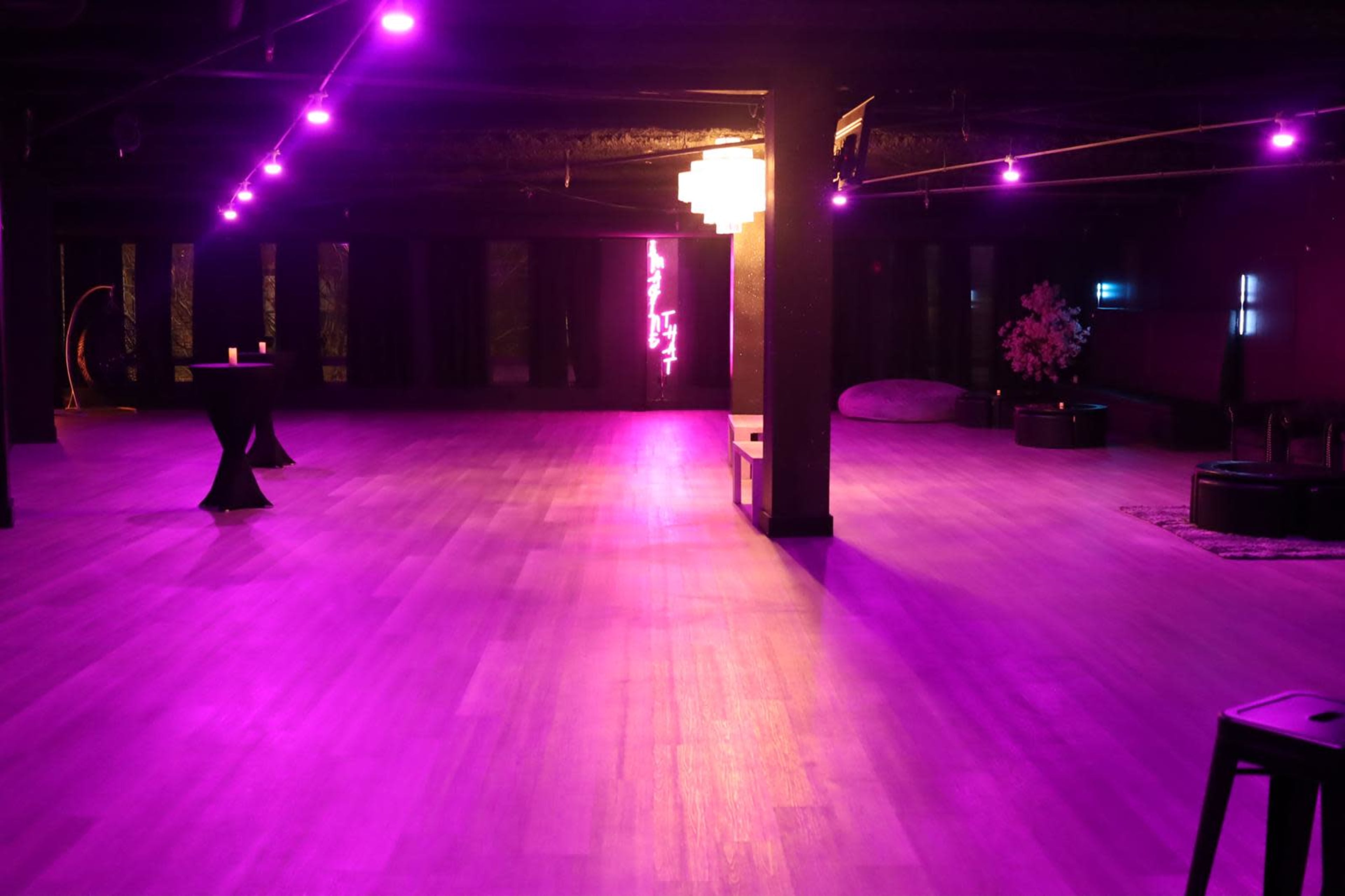 The image shows a spacious room with purple lighting, wooden flooring, and minimalist furniture arranged around.