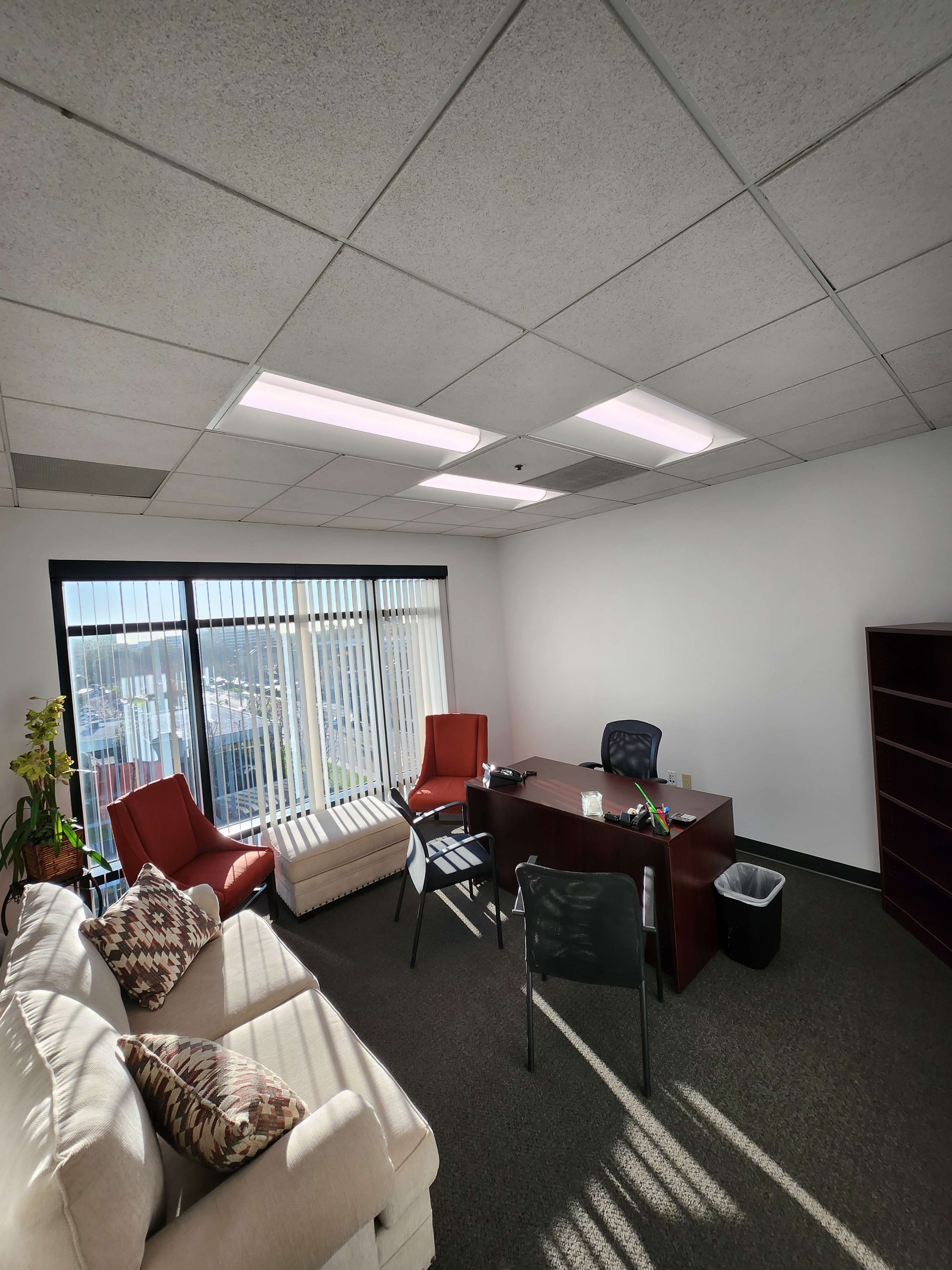 The image shows an office space with a desk, chairs, a couch, and large windows offering natural light.