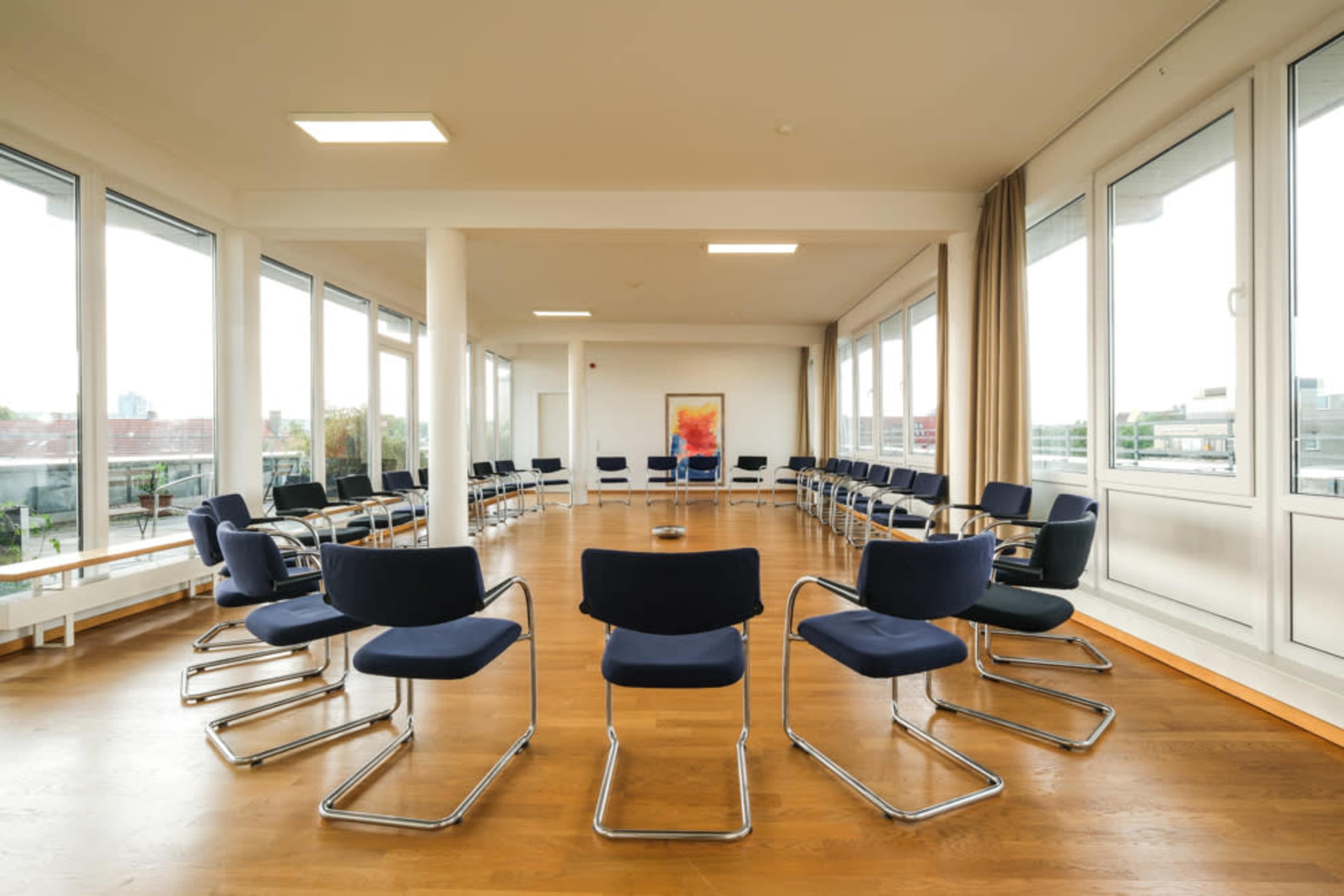 The image shows a spacious room arranged with multiple chairs in a circular formation around a central area, surrounded by large windows that allow natural light.