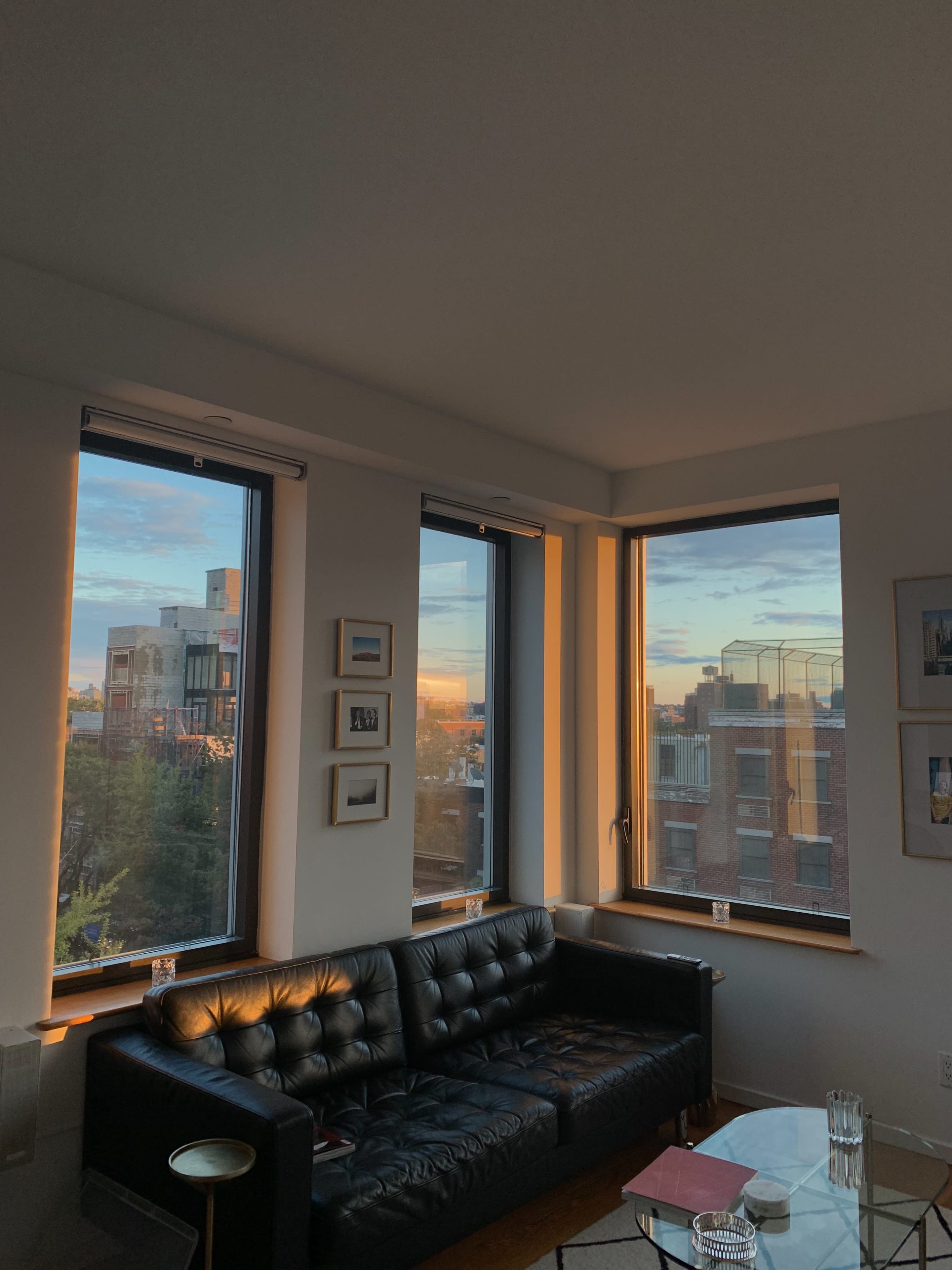 A modern living room with a black leather couch and large windows displaying a sunset over a cityscape.