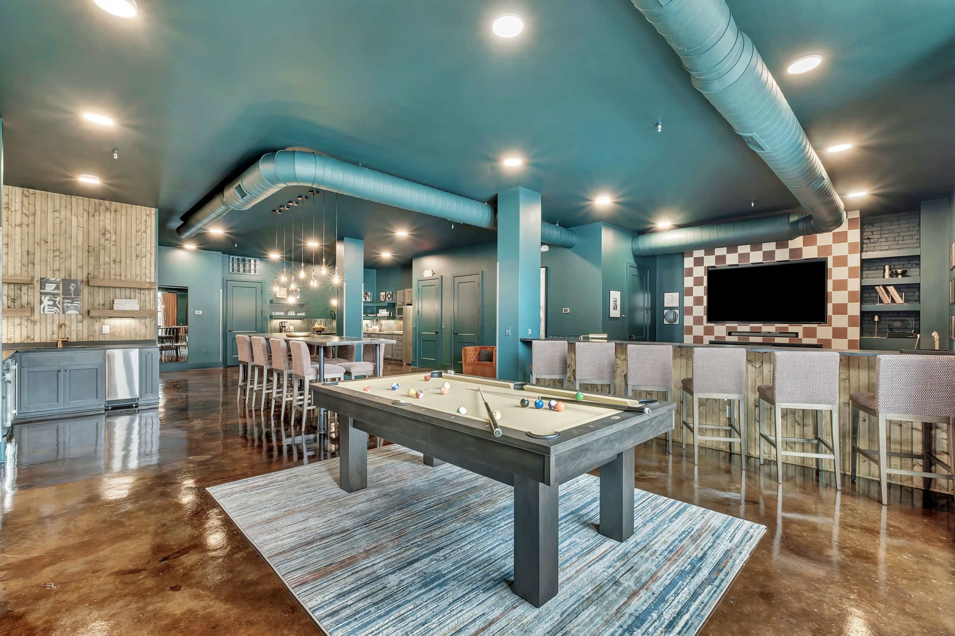 The image shows a modern lounge area featuring a pool table, a bar with seating, and a large television against a blue-green wall.