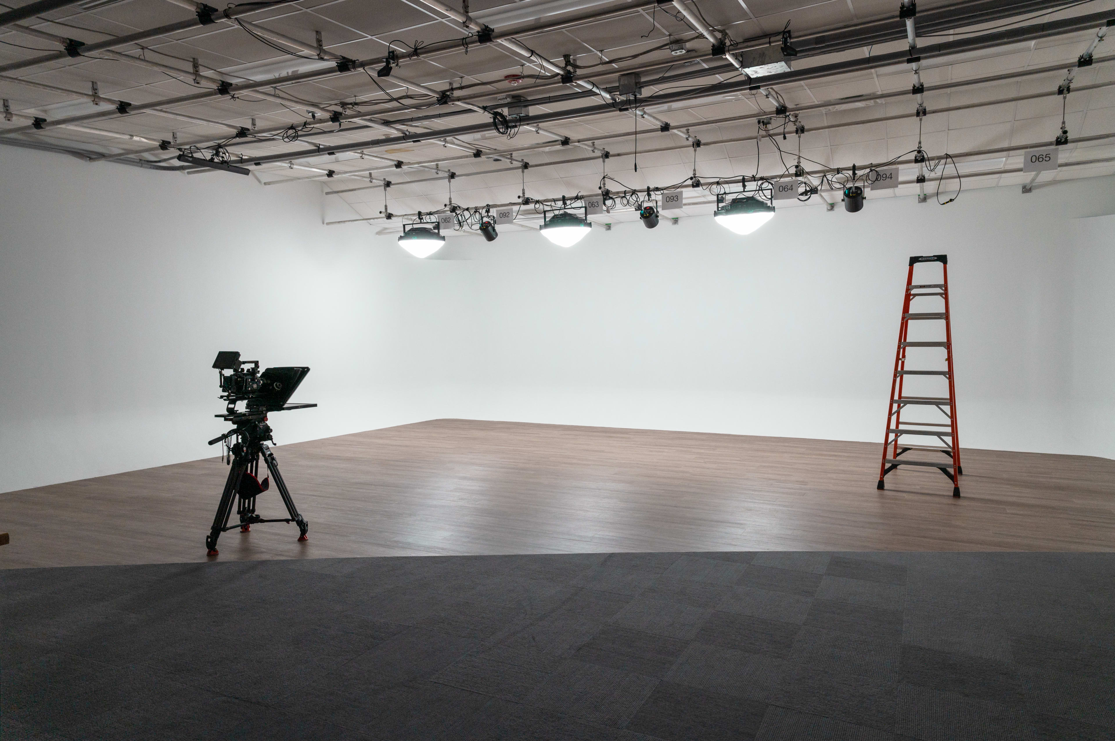 A minimalist studio space with a camera on a tripod, a ladder, and overhead lighting fixtures.