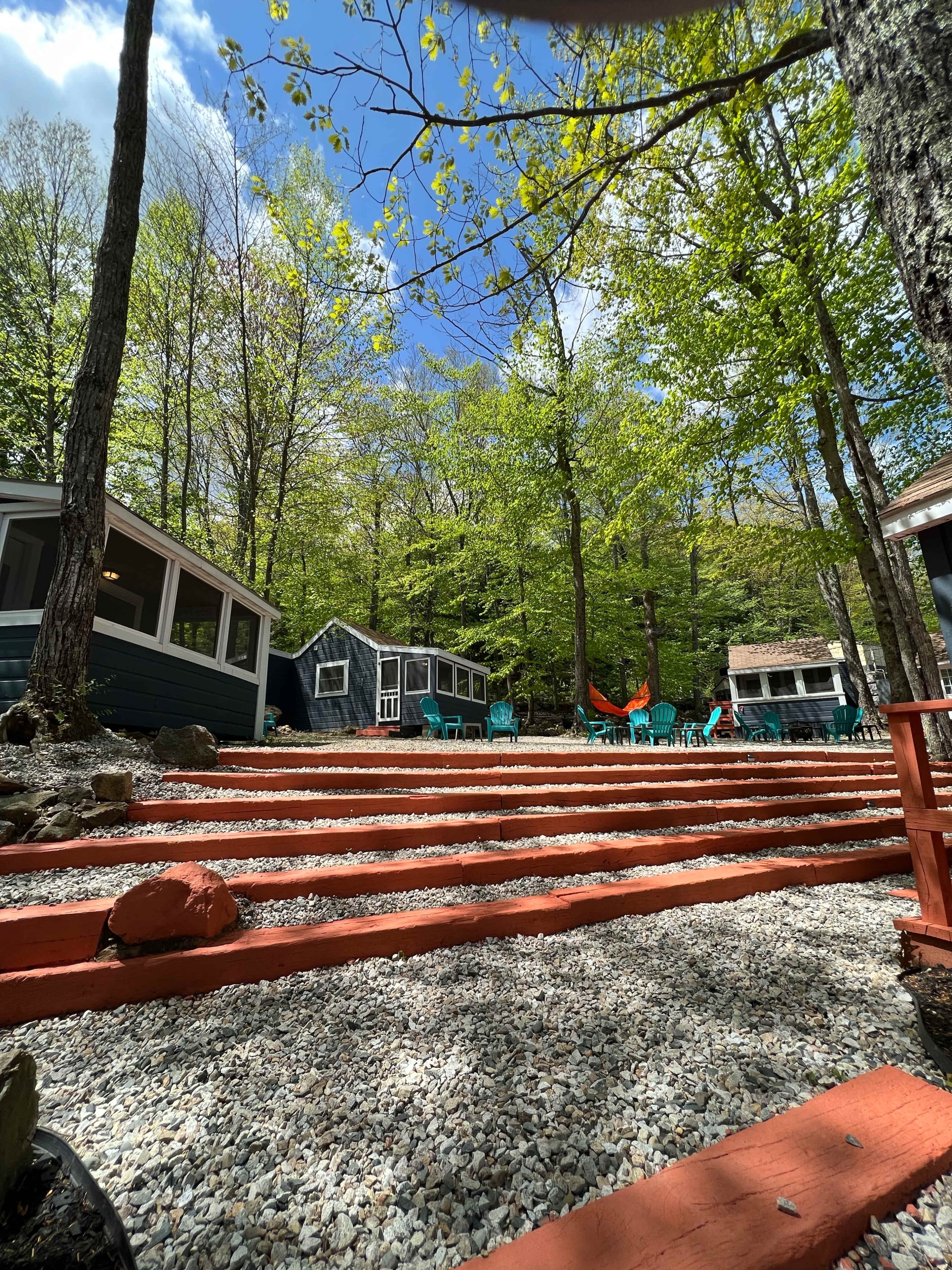 A wooded area with a series of cabins, stone steps, and turquoise chairs surrounded by trees under a clear blue sky.