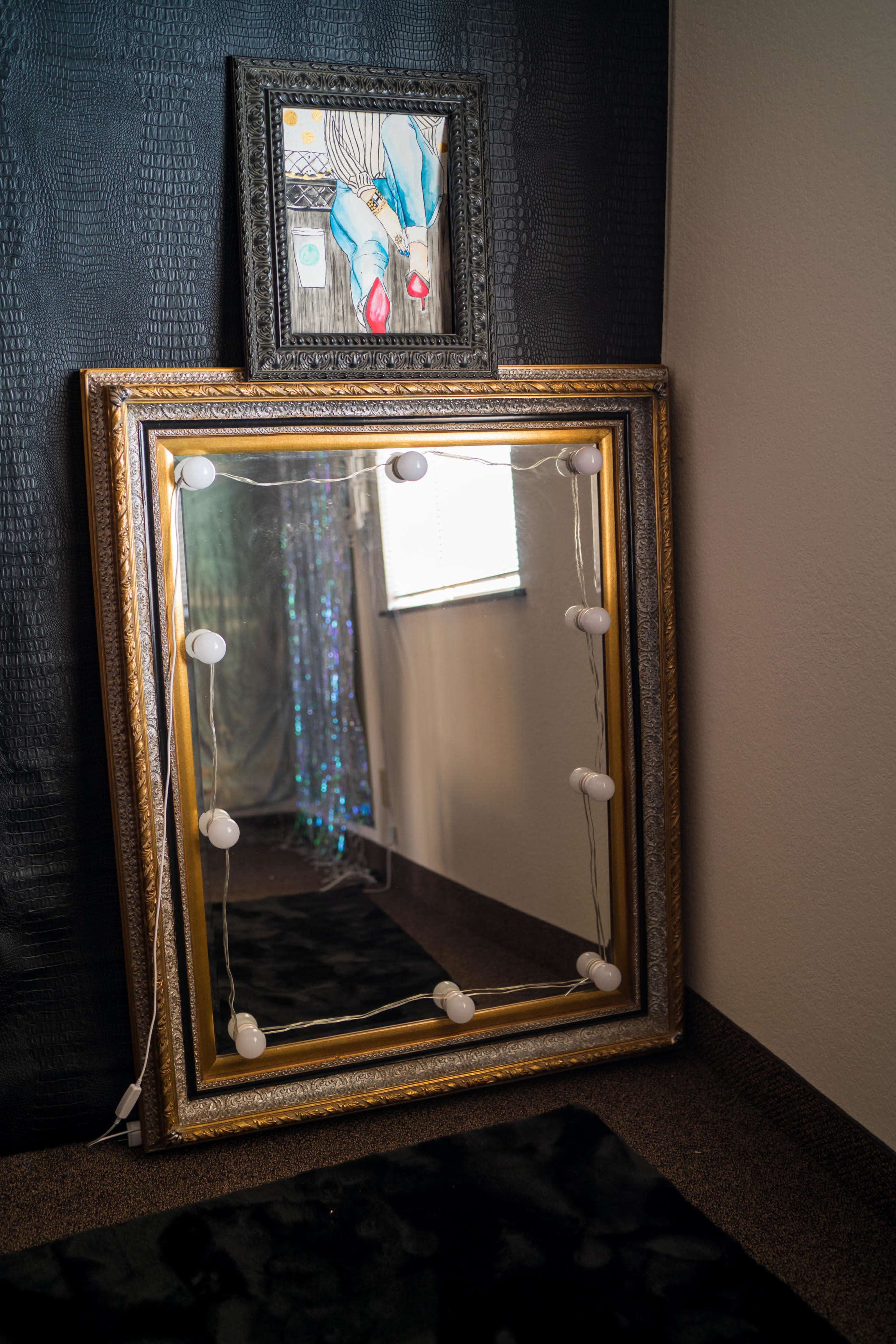 A large ornate mirror with lights around its frame stands against a wall beneath a framed artwork.
