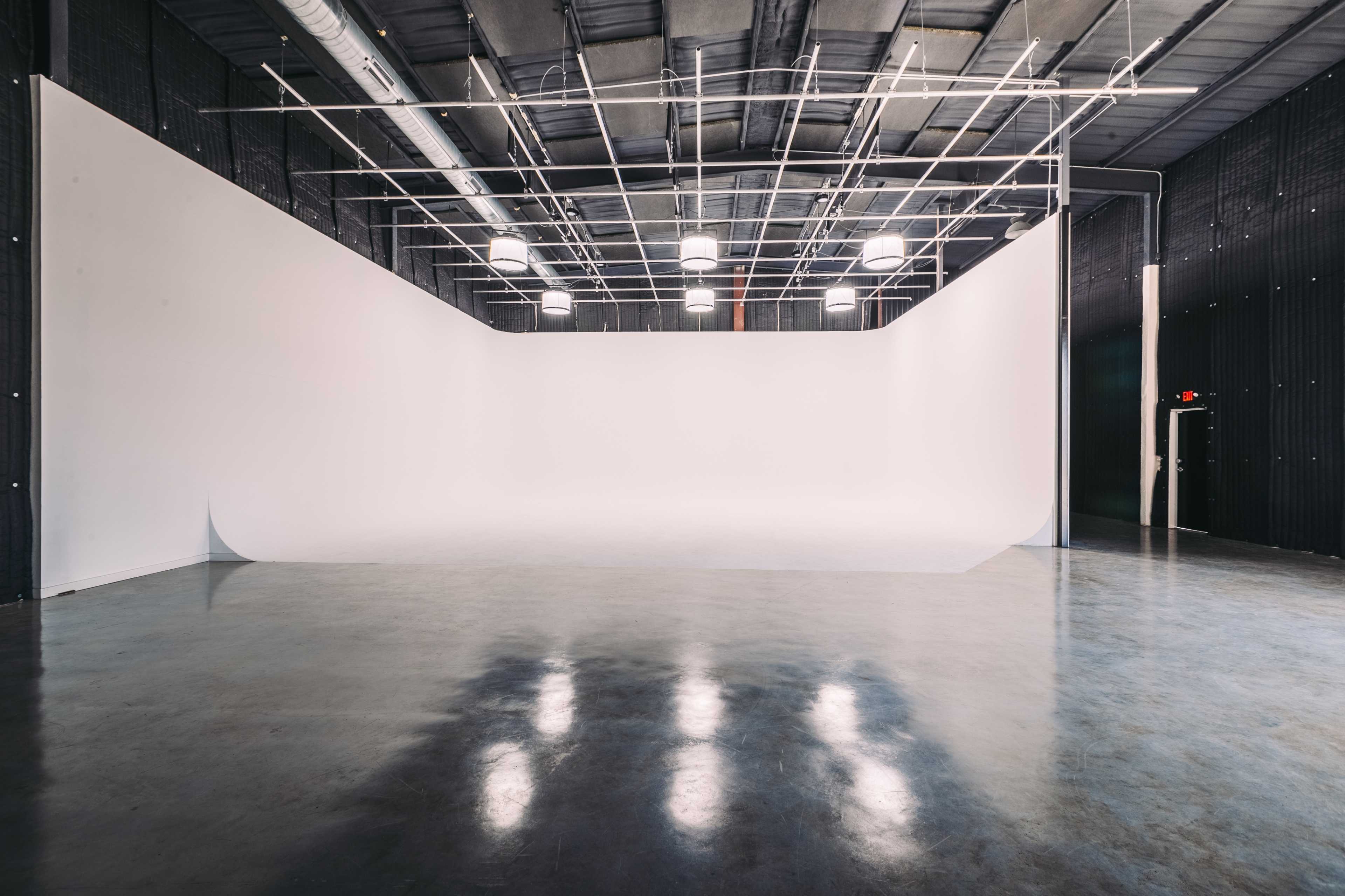 A spacious, empty photography studio with a white backdrop and overhead lighting.