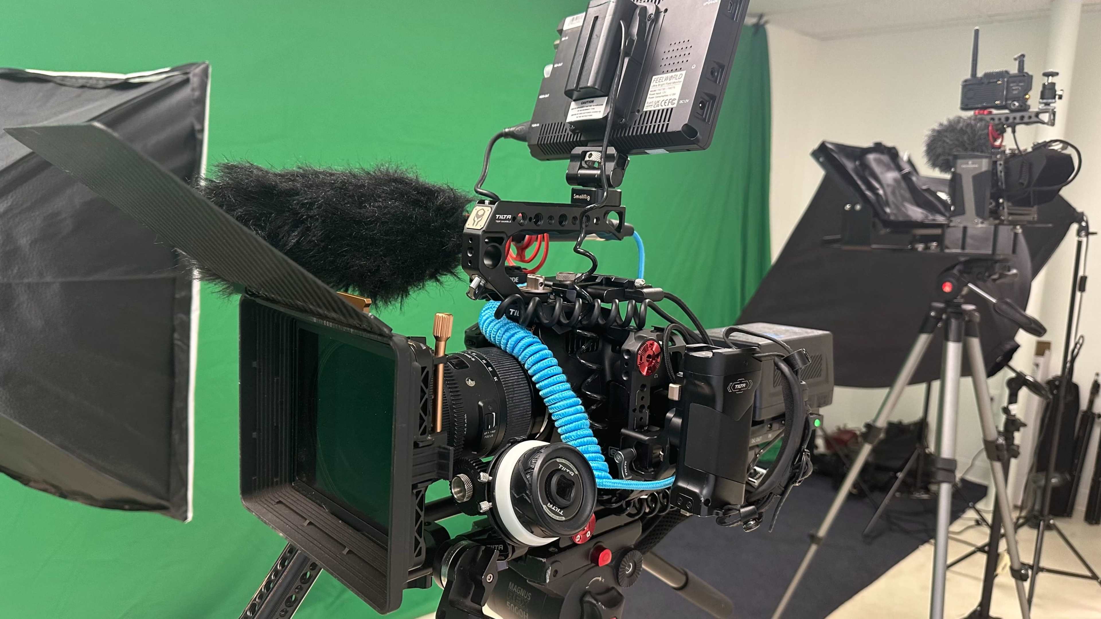 A camera setup with a green screen backdrop, featuring professional video equipment including microphones and monitors on tripods.