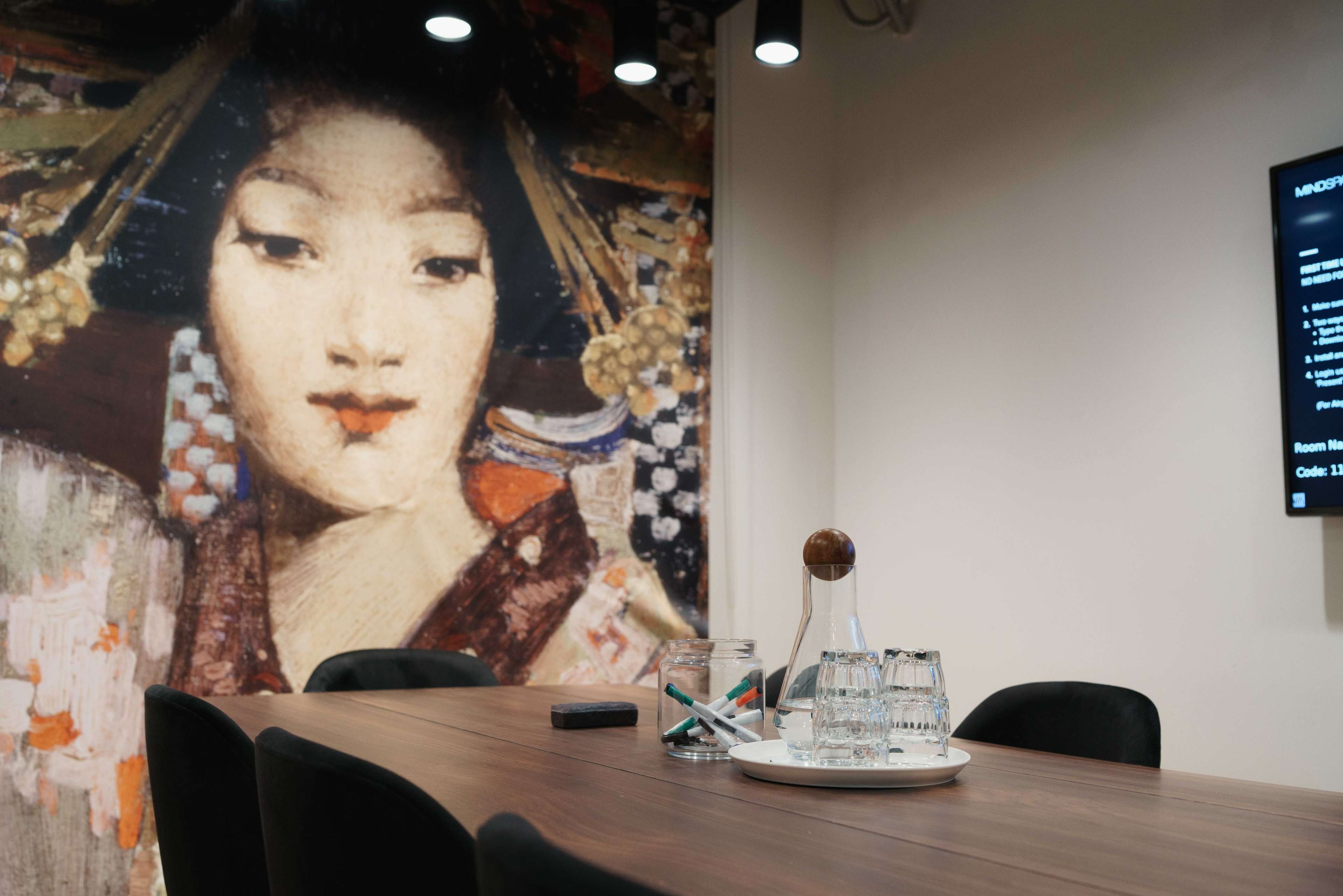 A conference room features a wooden table with glasses and a glass decanter, set against a mural of a woman in traditional attire.