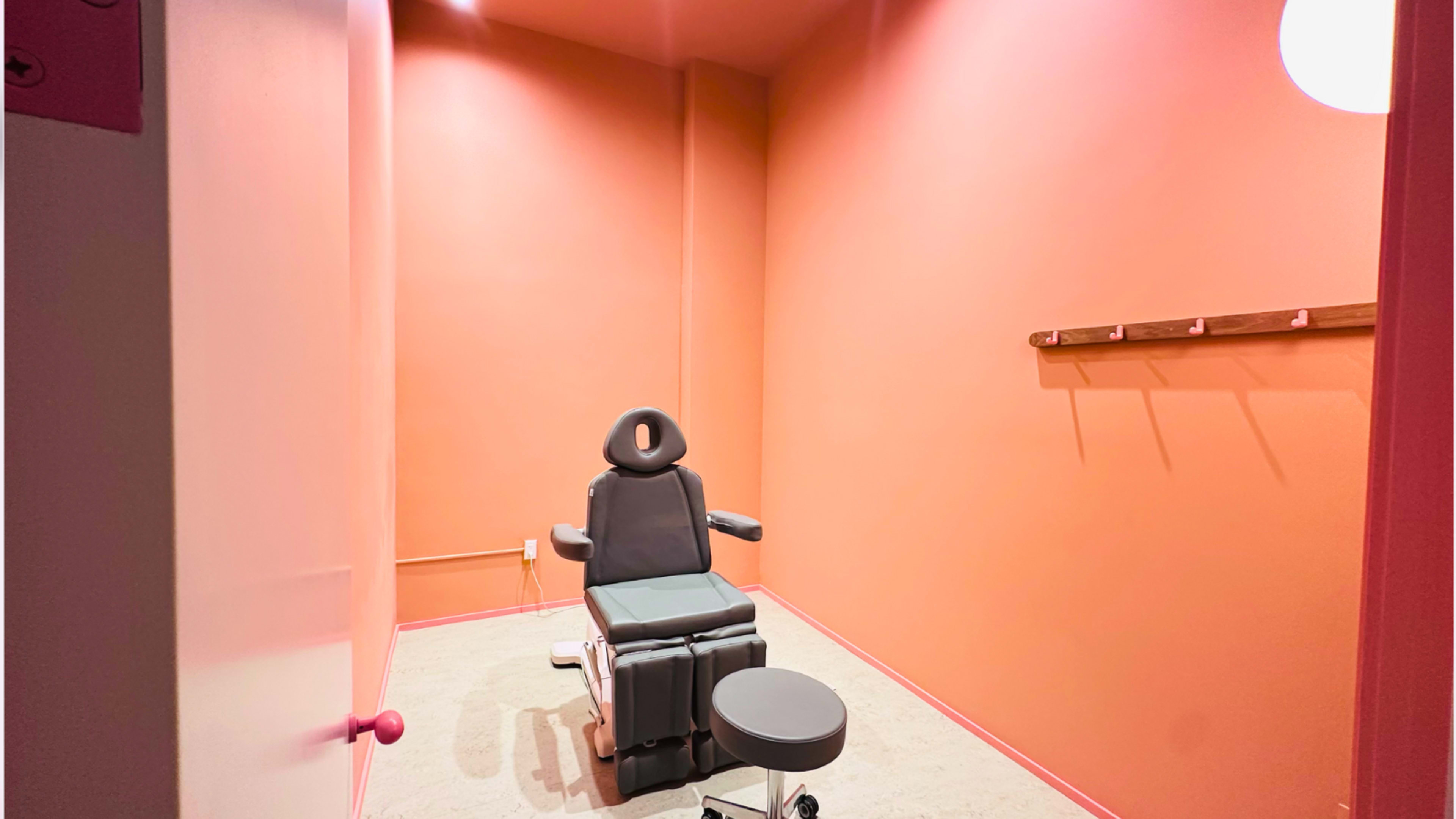 The image shows a small, empty treatment room with an examination chair and a stool on a smooth floor, all set against orange walls.