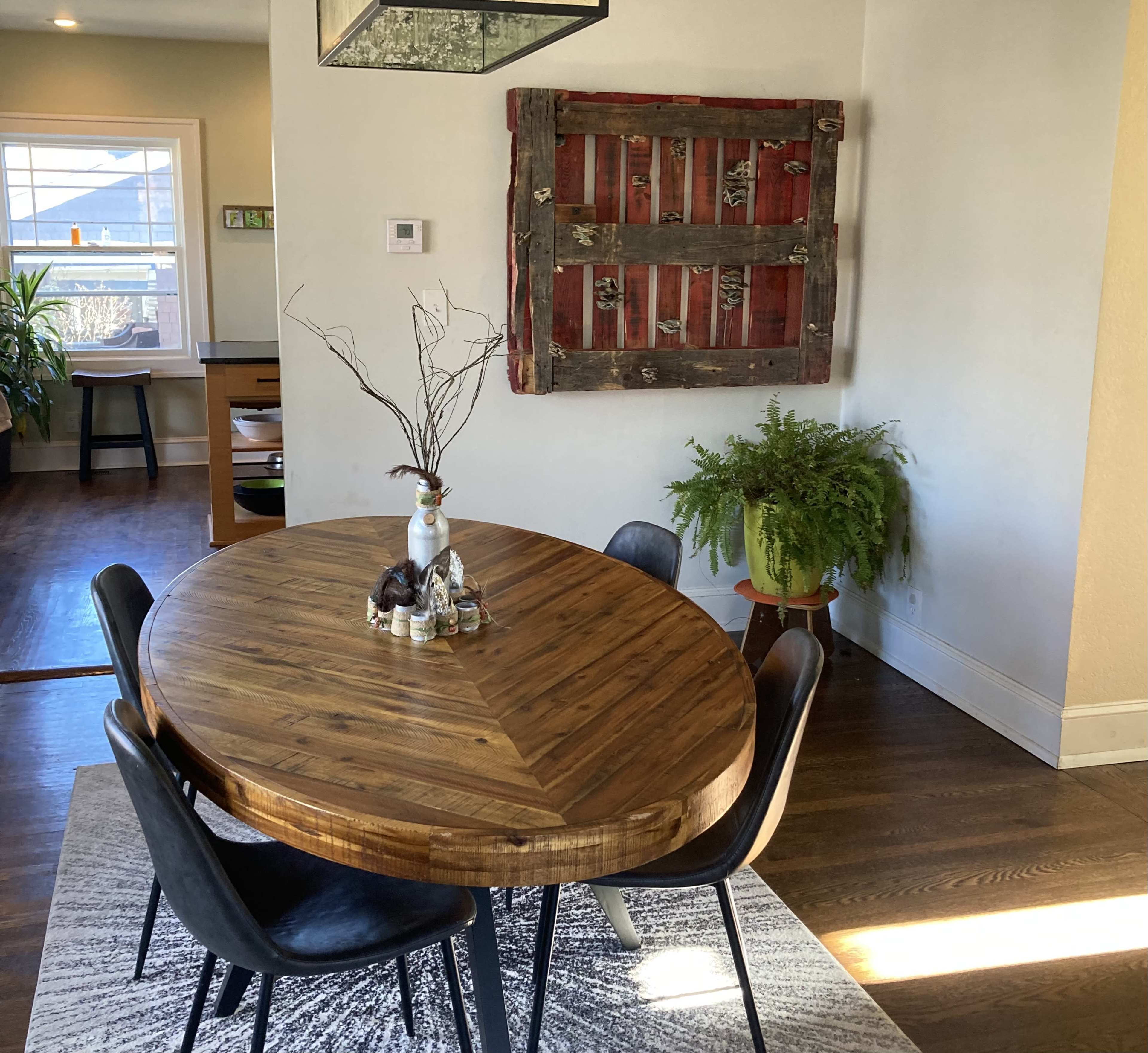 A round wooden table is set with a vase and decorations, surrounded by black chairs, with a red and wooden wall art nearby and a plant in a pot.