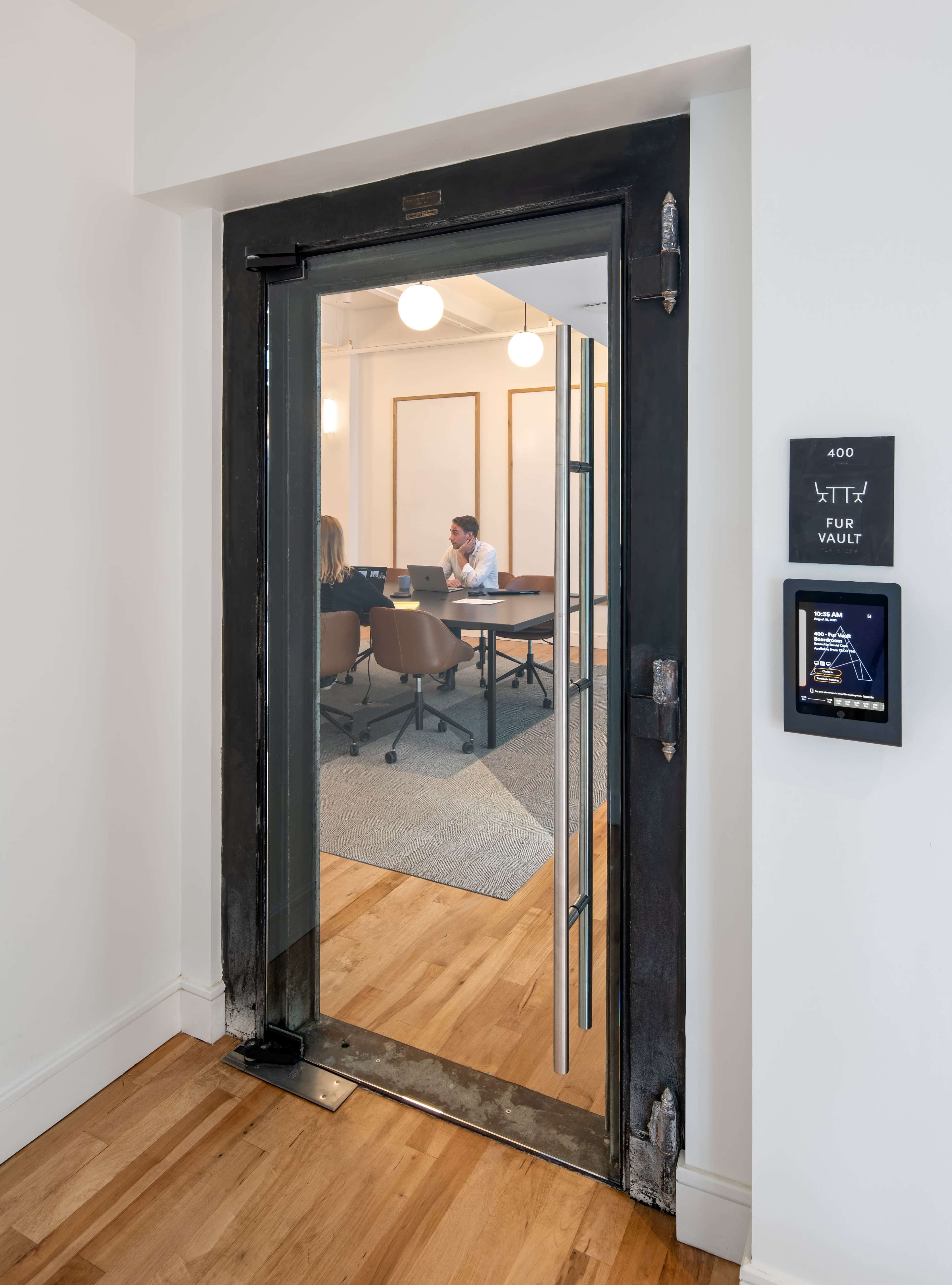 A glass and metal door opens to a meeting room with a table and chairs, where two people are engaged in conversation.