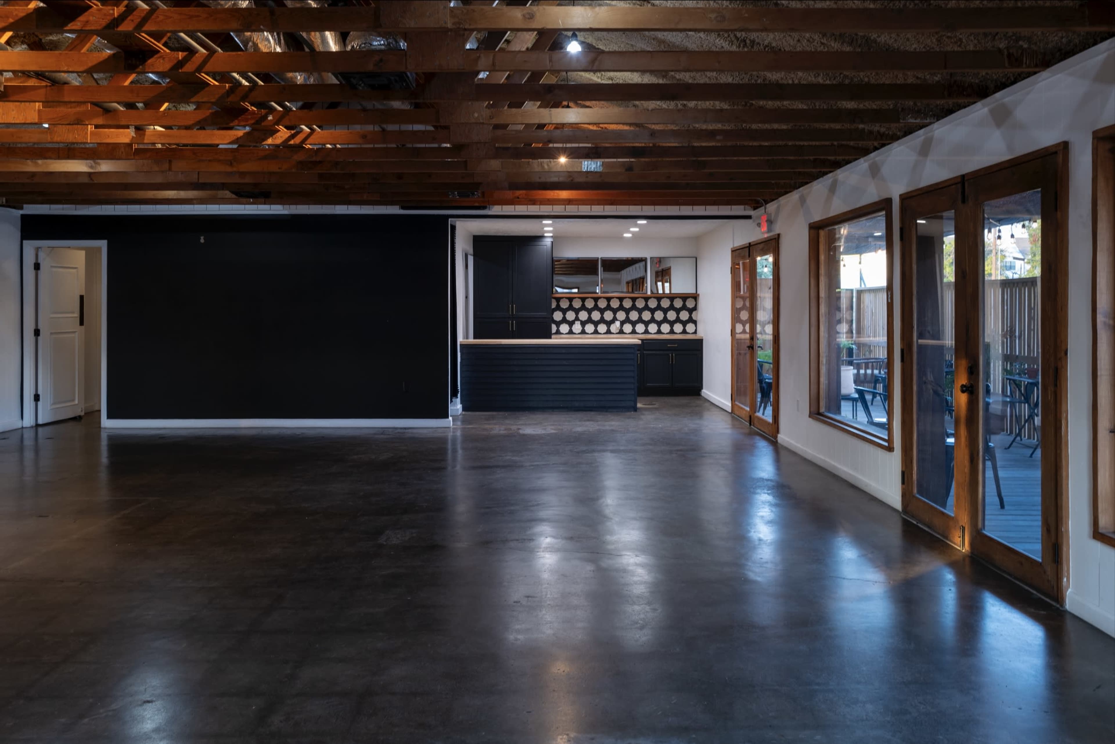 An interior space with polished concrete floors, exposed wooden beams, and large windows, featuring a black wall and a bar area in the background.