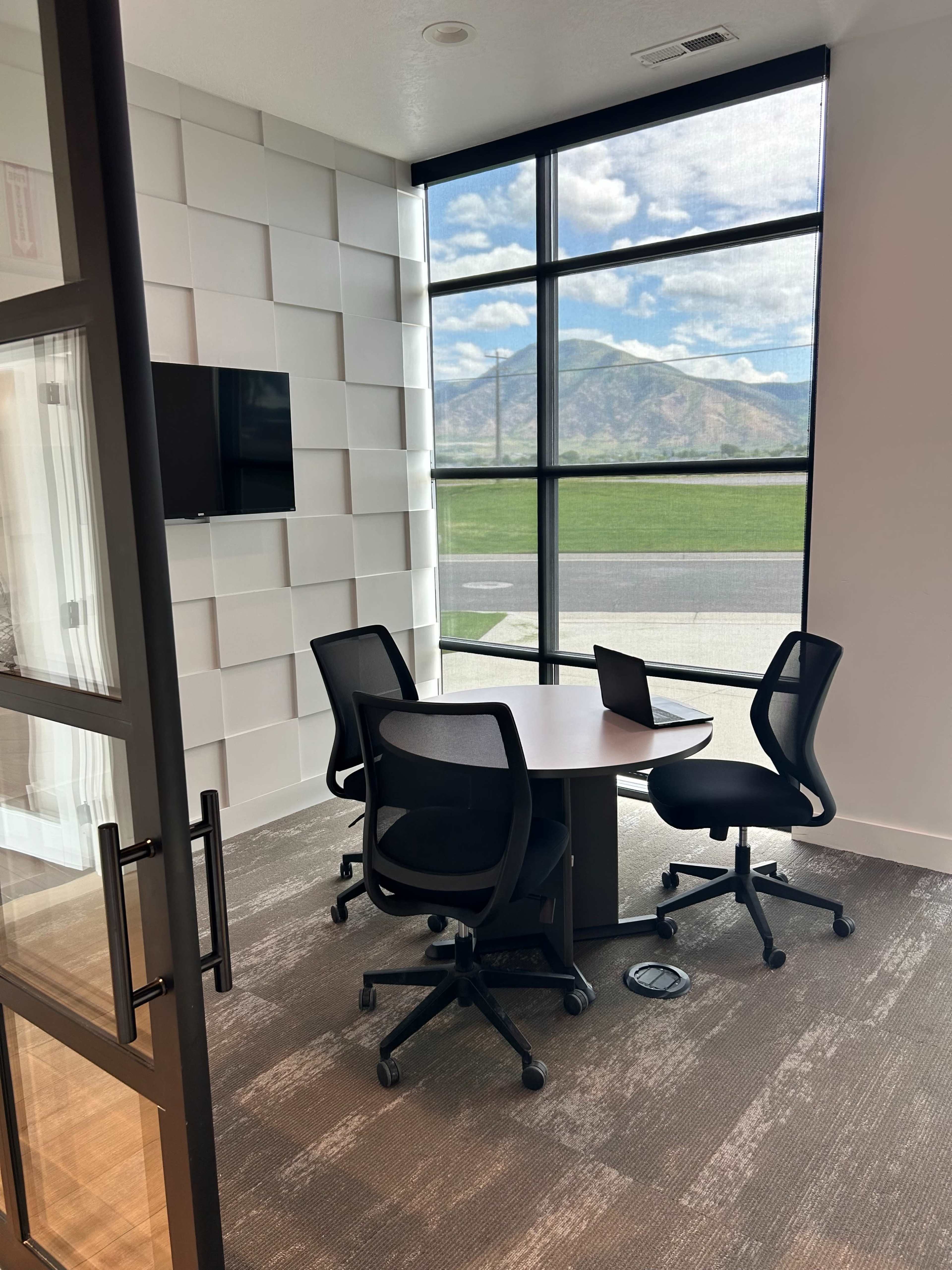 A small conference room features a round table with four chairs, a wall-mounted TV, and large windows showcasing a mountain view.