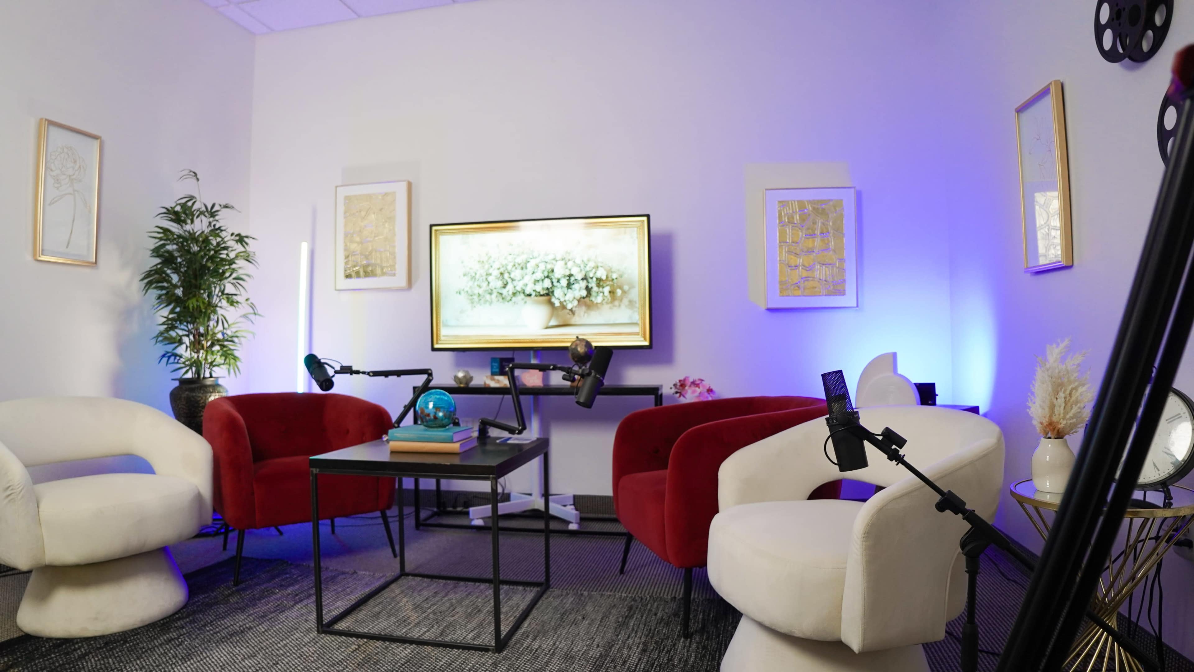 The image shows a modern indoor space with two red chairs and one white chair arranged around a black coffee table, a television displaying flowers on the wall, and ambient lighting.