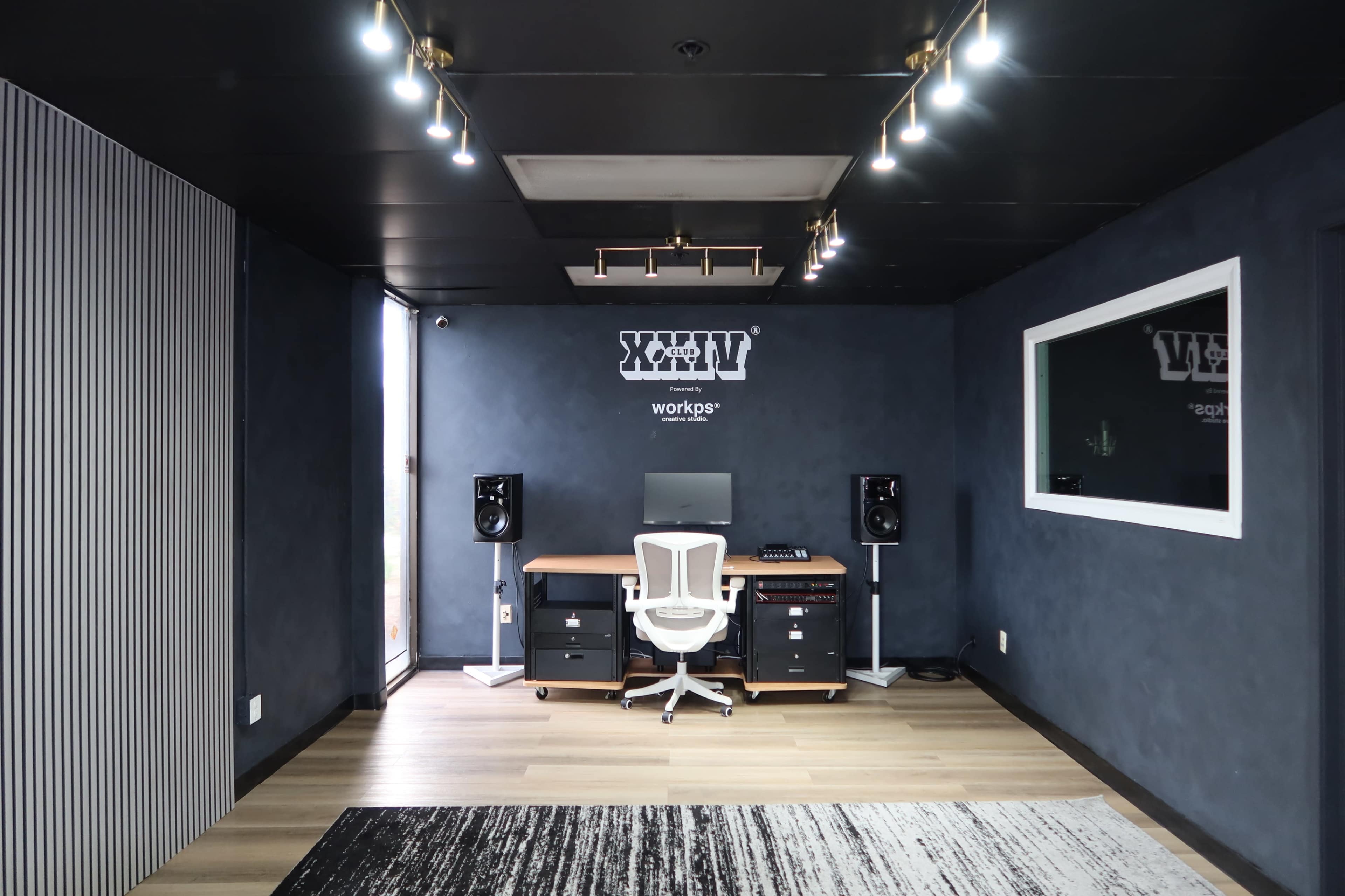 The image shows a modern recording studio with a desk, computer, and audio equipment illuminated by ceiling lights, featuring dark walls and wooden flooring.