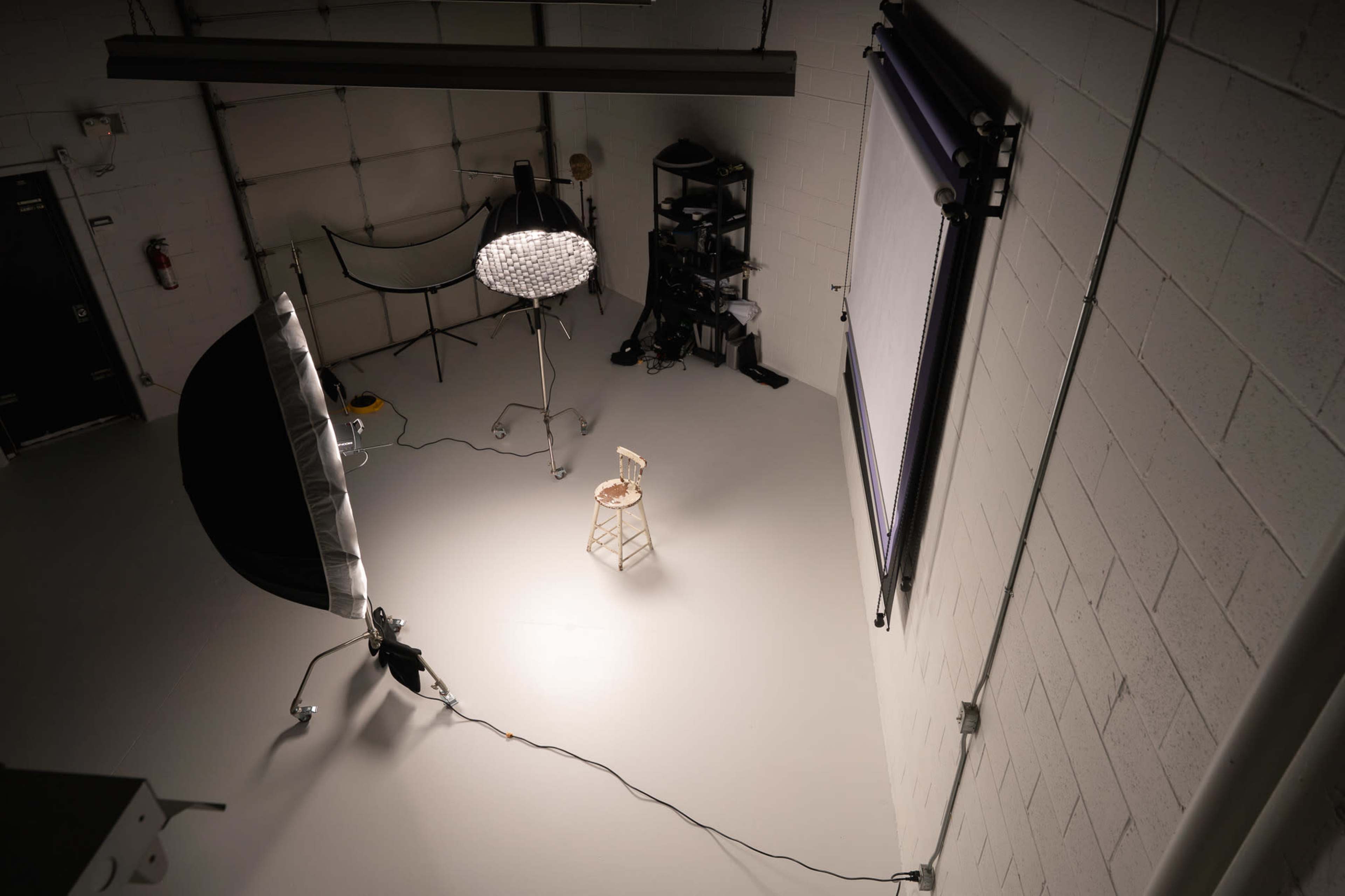 The image shows a photography studio with a stool positioned in the center, illuminated by a light source, and equipped with various lighting setups and equipment in the background.