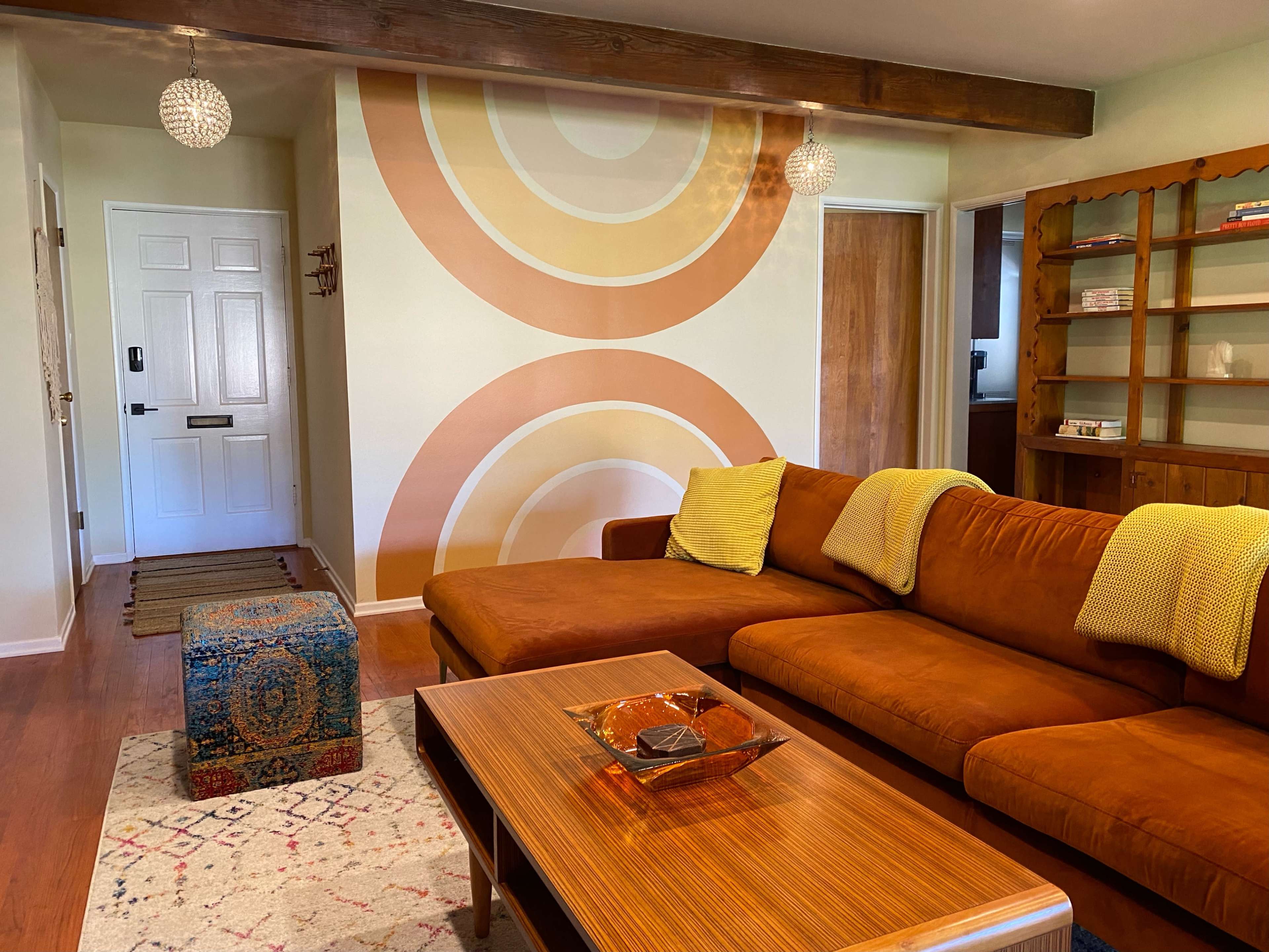 The living room features a brown sectional sofa with yellow throw pillows, a wooden coffee table, a patterned rug, and a retro wall mural of concentric circles.