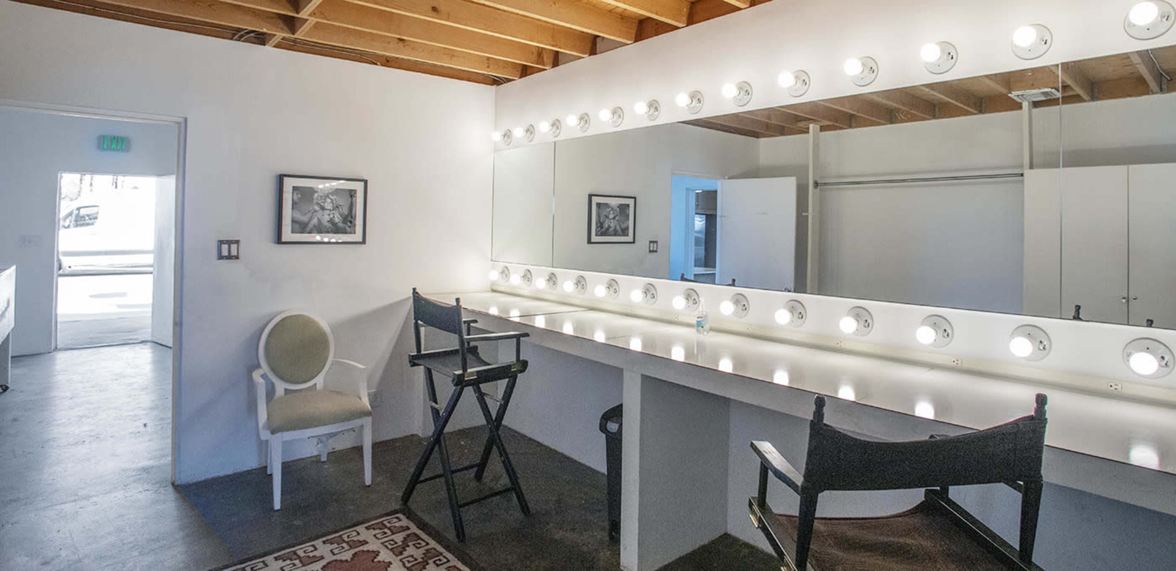 A well-lit dressing room with a large mirror surrounded by lights, two director's chairs, and a white sofa across from the entrance.