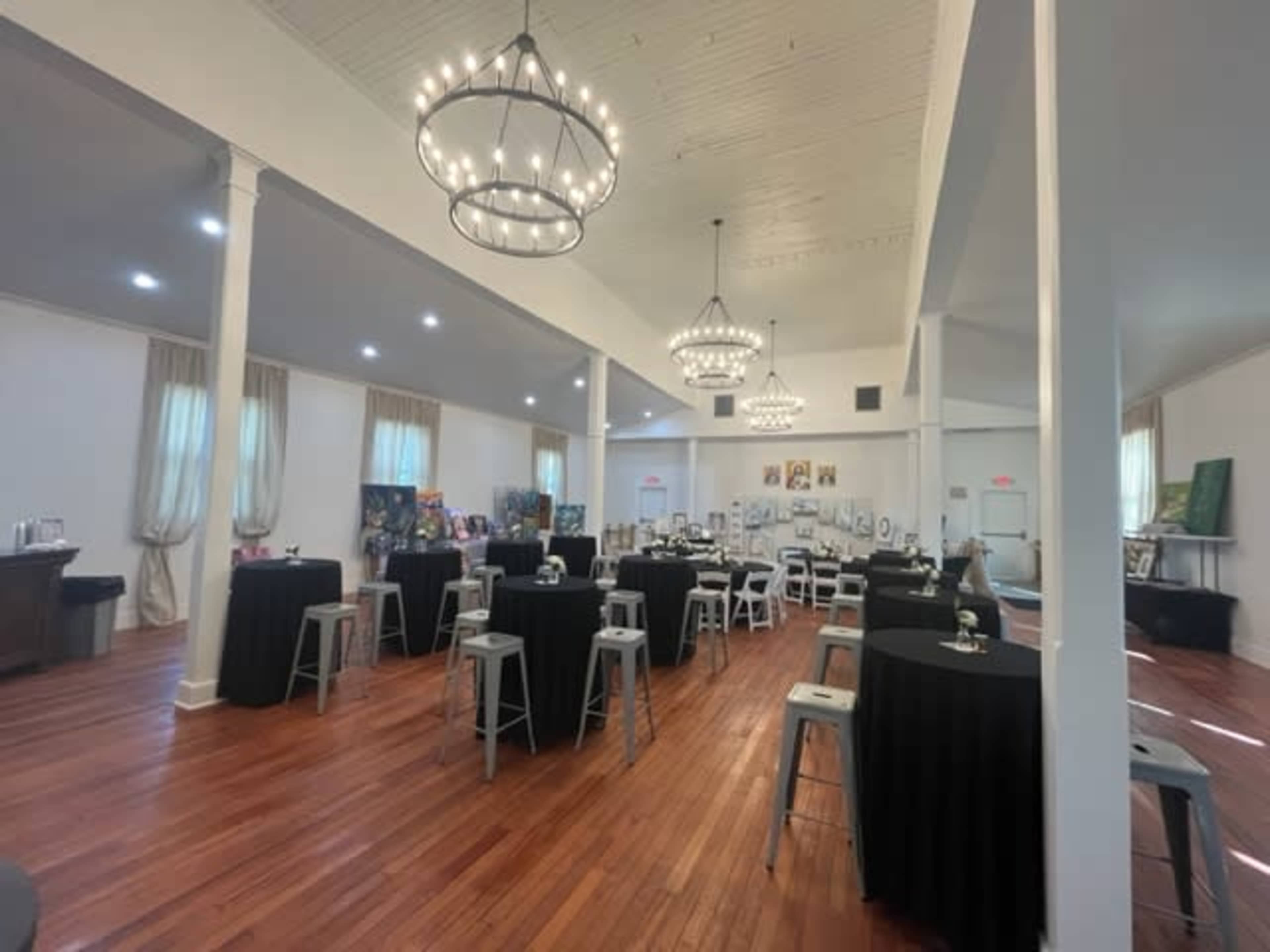 An elegantly arranged event space with several black tablecloth-covered tables, wooden flooring, and chandeliers hanging from a white ceiling.
