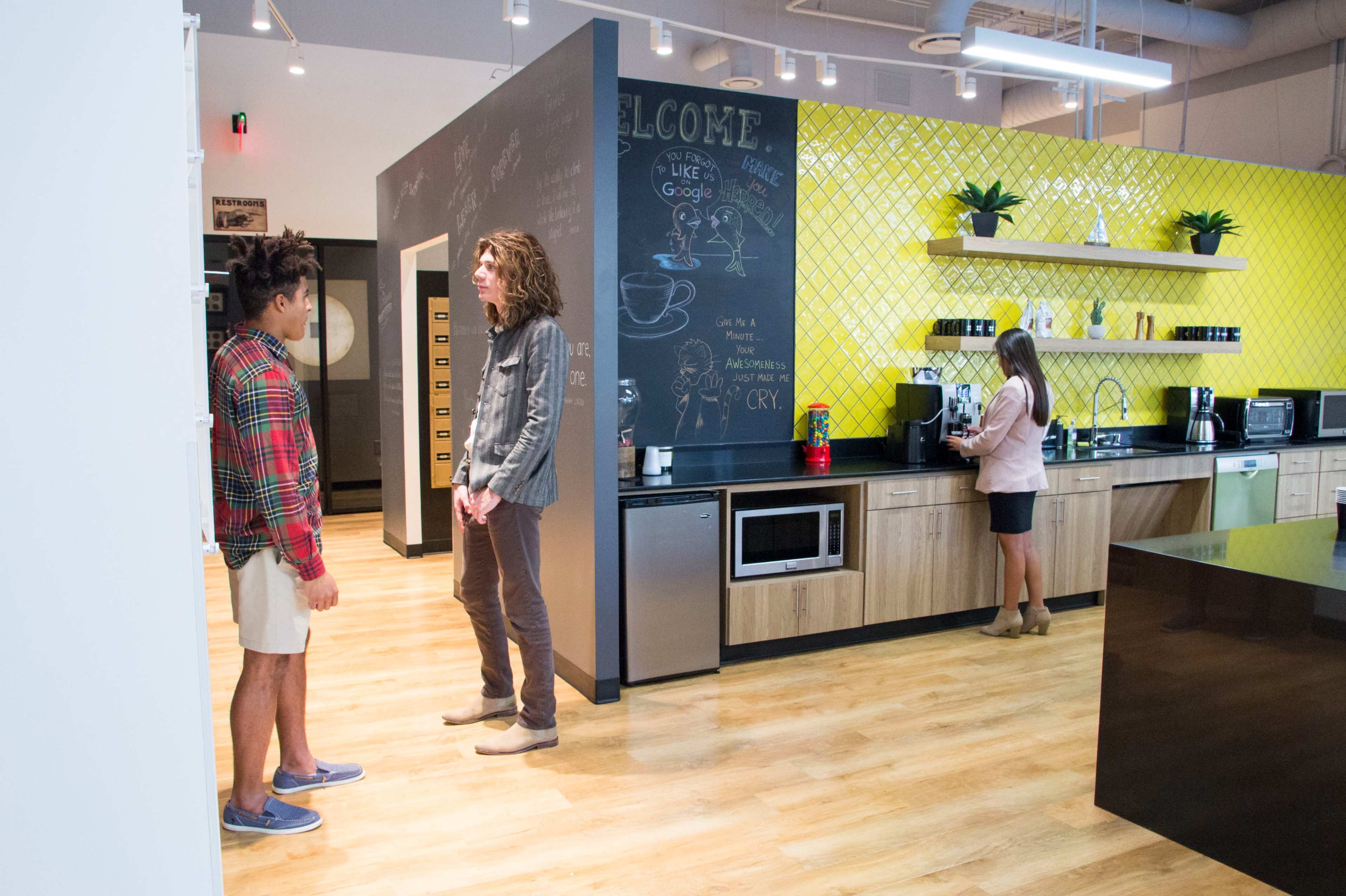 Two people converse in a modern kitchen area with wood flooring and a colorful accent wall featuring a chalkboard, while a third person prepares coffee at a counter.