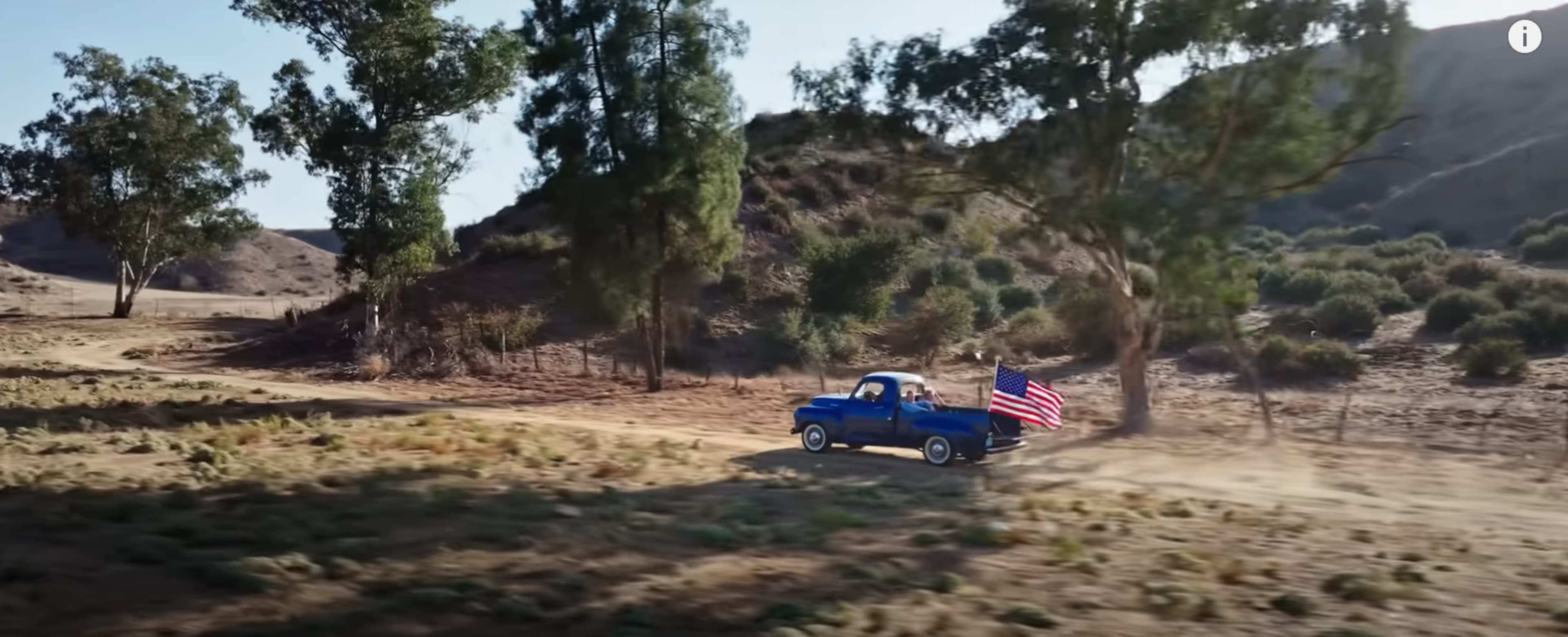 A blue pickup truck drives along a dirt road in a rural area, displaying an American flag.