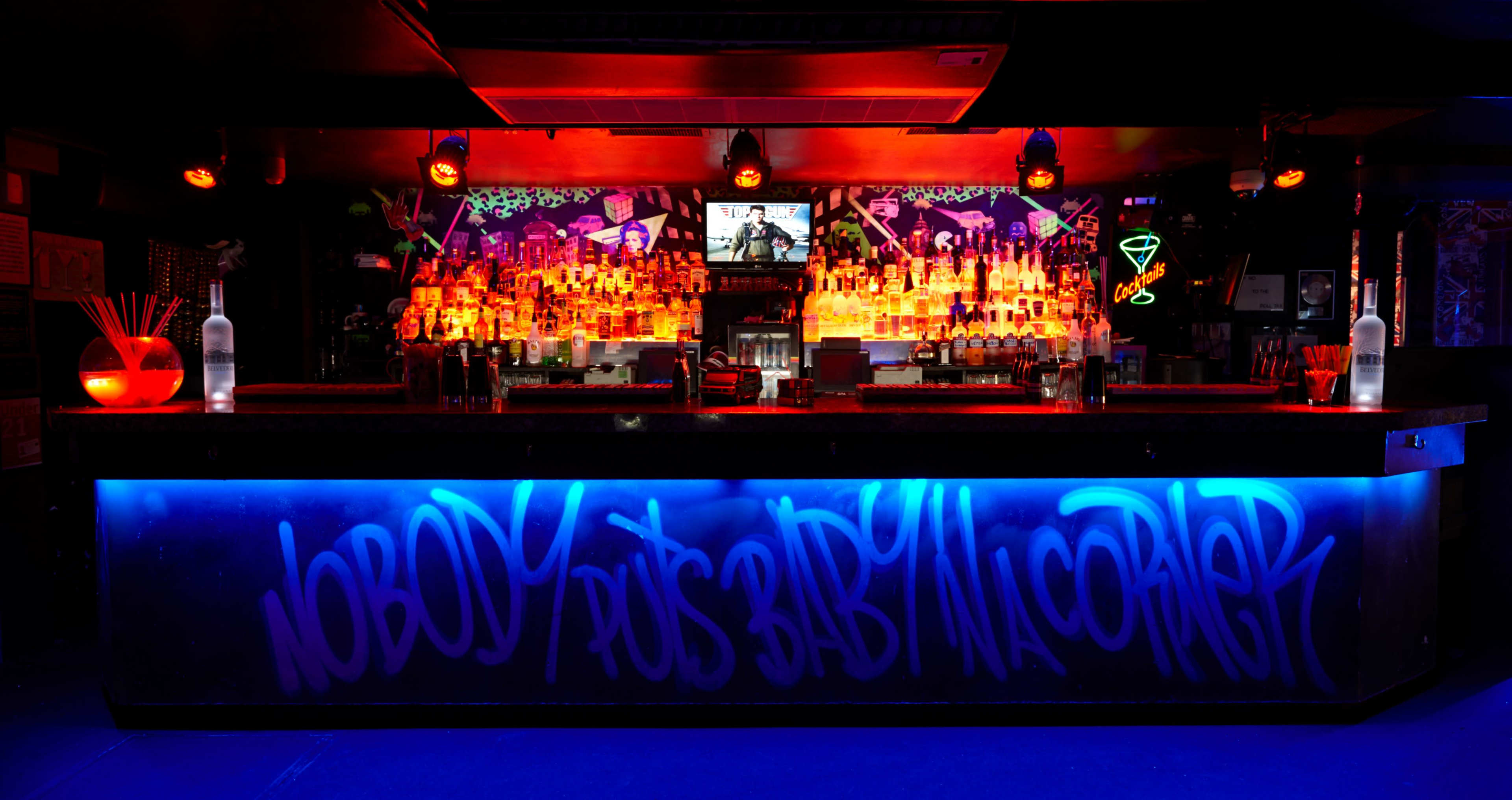 A brightly lit bar counter with a colorful array of bottles and neon decorations features graffiti on the front that reads, "NOBODY PUTS BABY IN A CORNER."