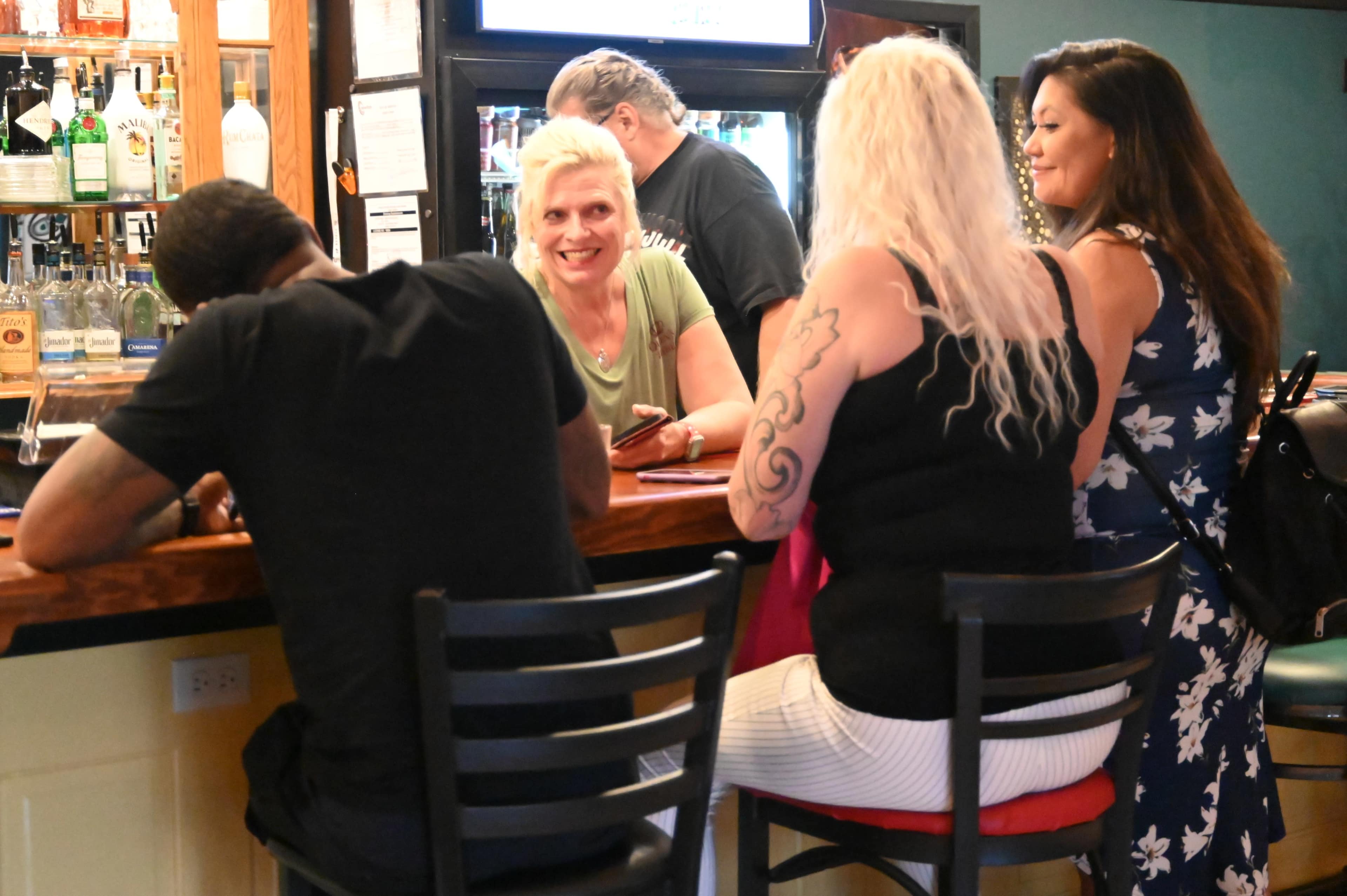 A group of people are socializing at a bar, with one bartender smiling at patrons and others engaged in conversation.