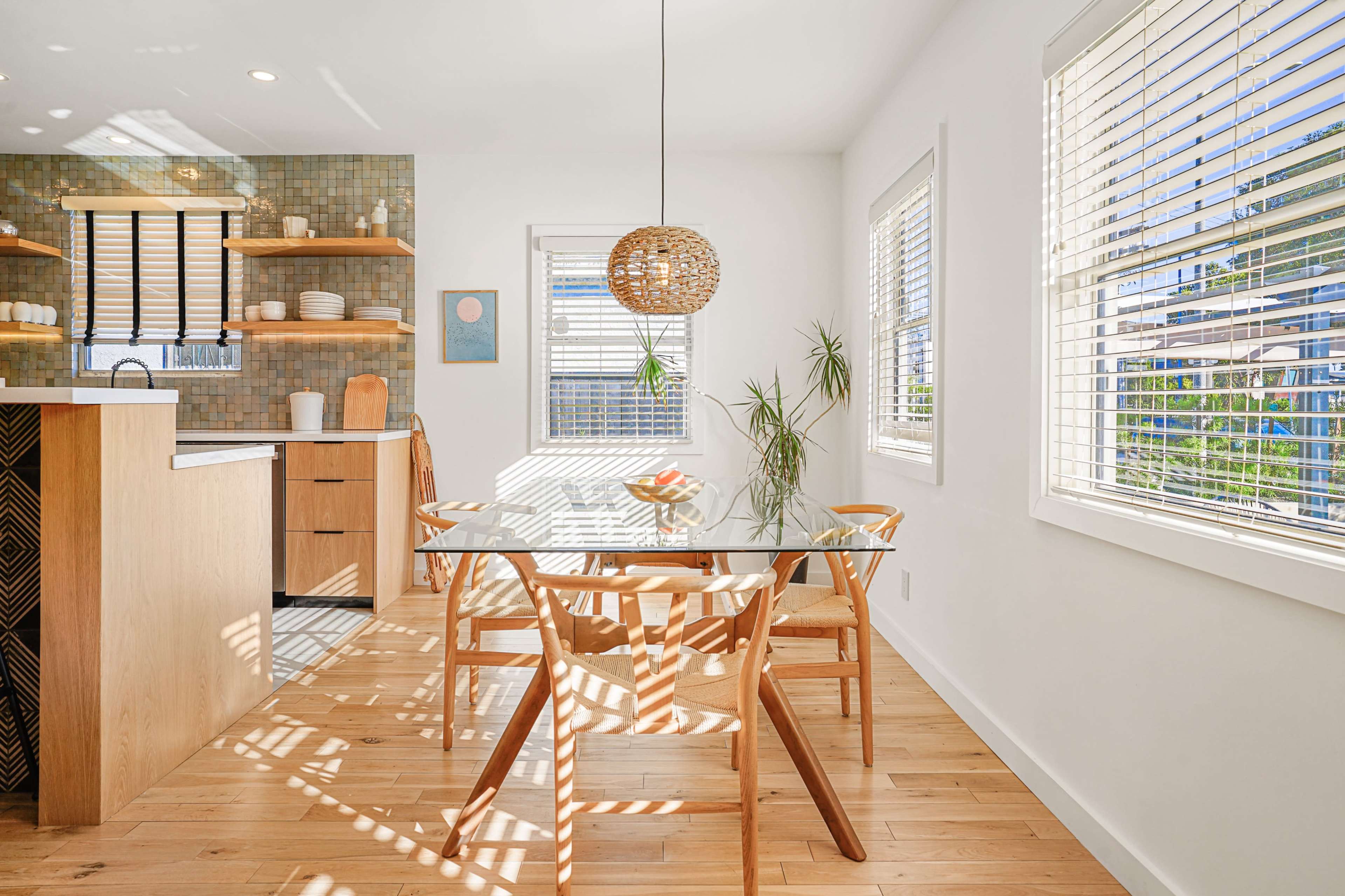 A bright dining area features a glass table surrounded by wooden chairs, with a modern kitchen in the background and sunlight streaming through the blinds.