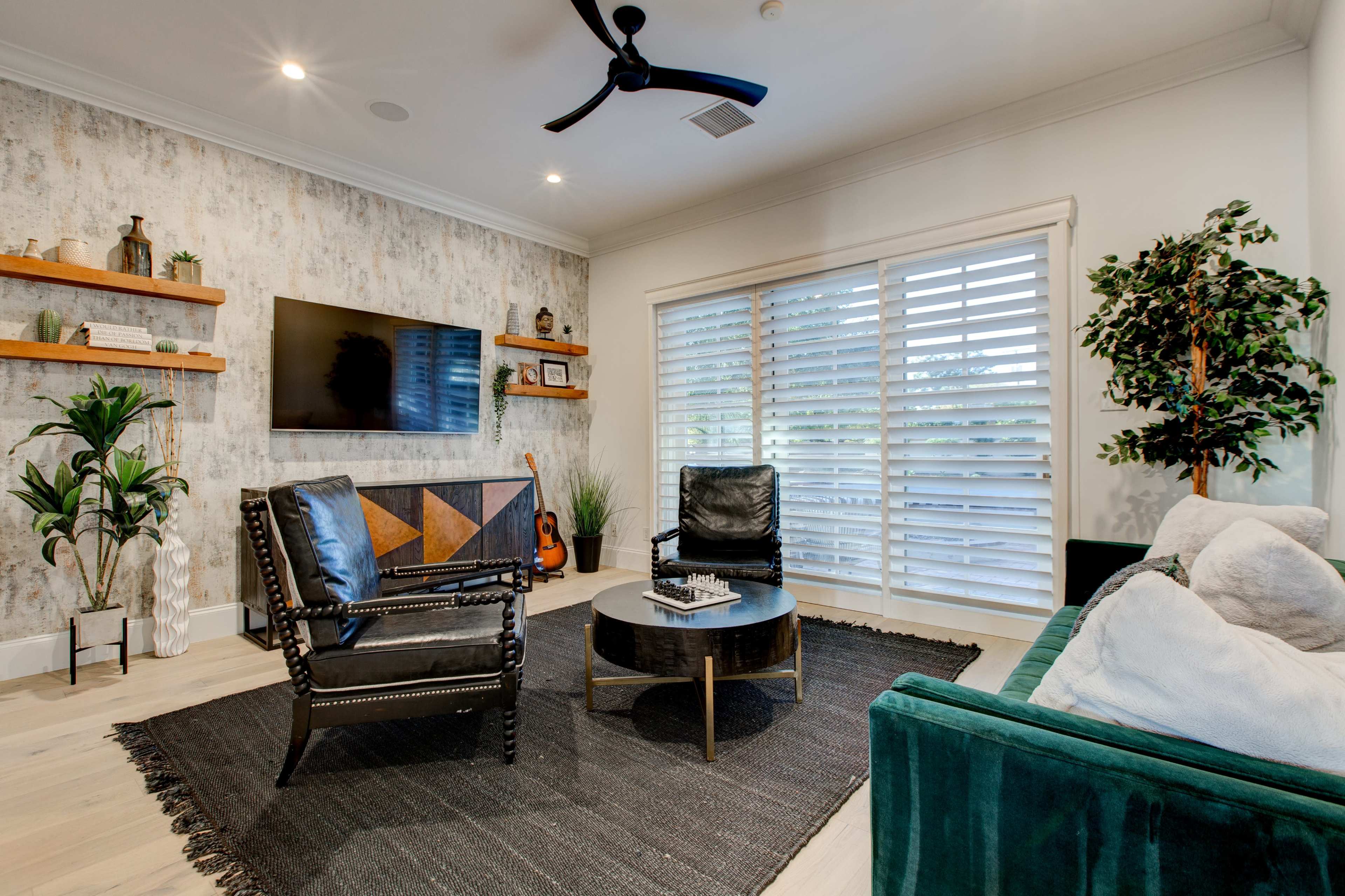 A modern living room features black chairs, a green sofa, a round coffee table, wall-mounted shelves, and a television, with natural light filtering through plantation shutters.