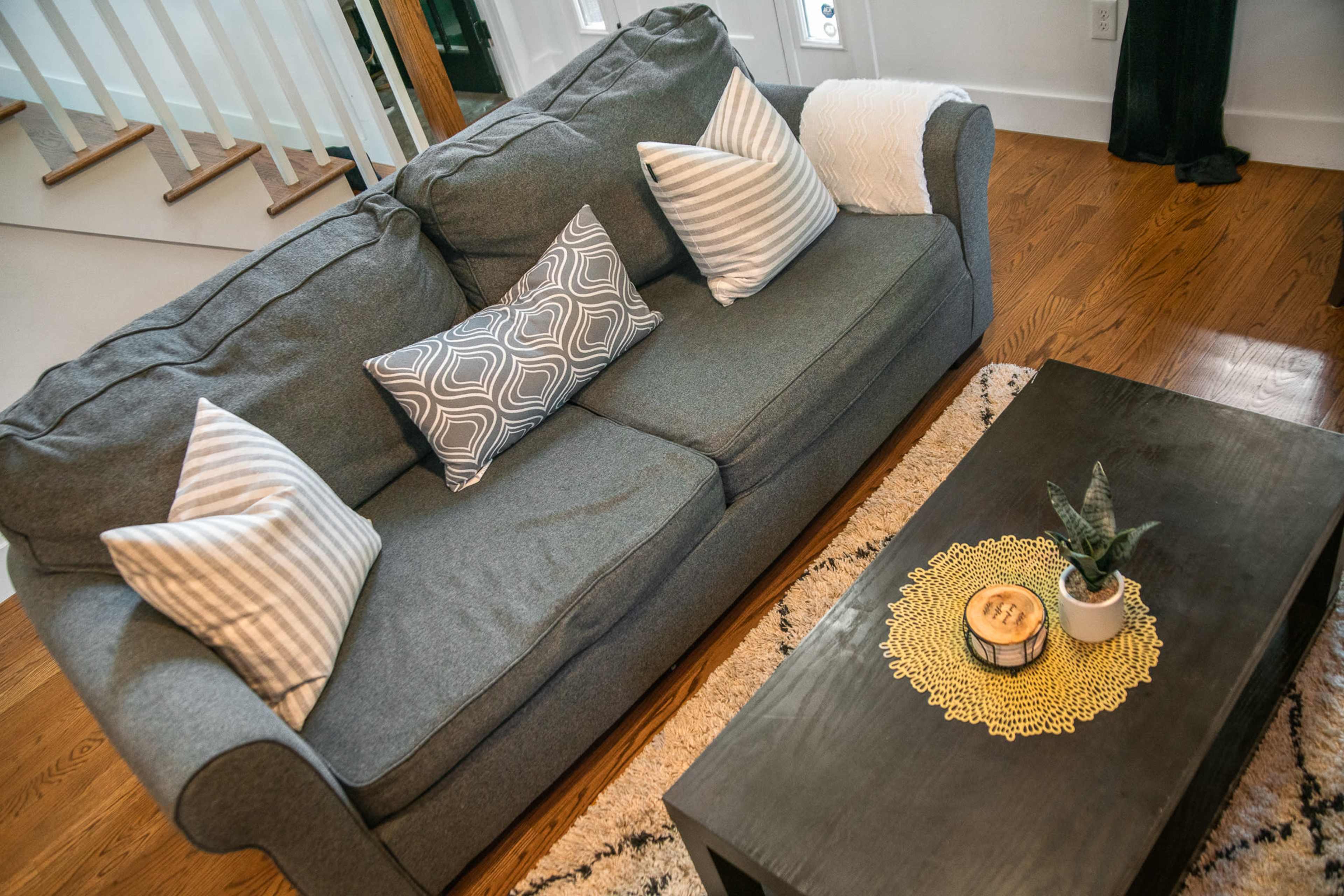 A gray sofa with patterned pillows sits in a living room, accompanied by a dark coffee table featuring a plant and a decorative tray.