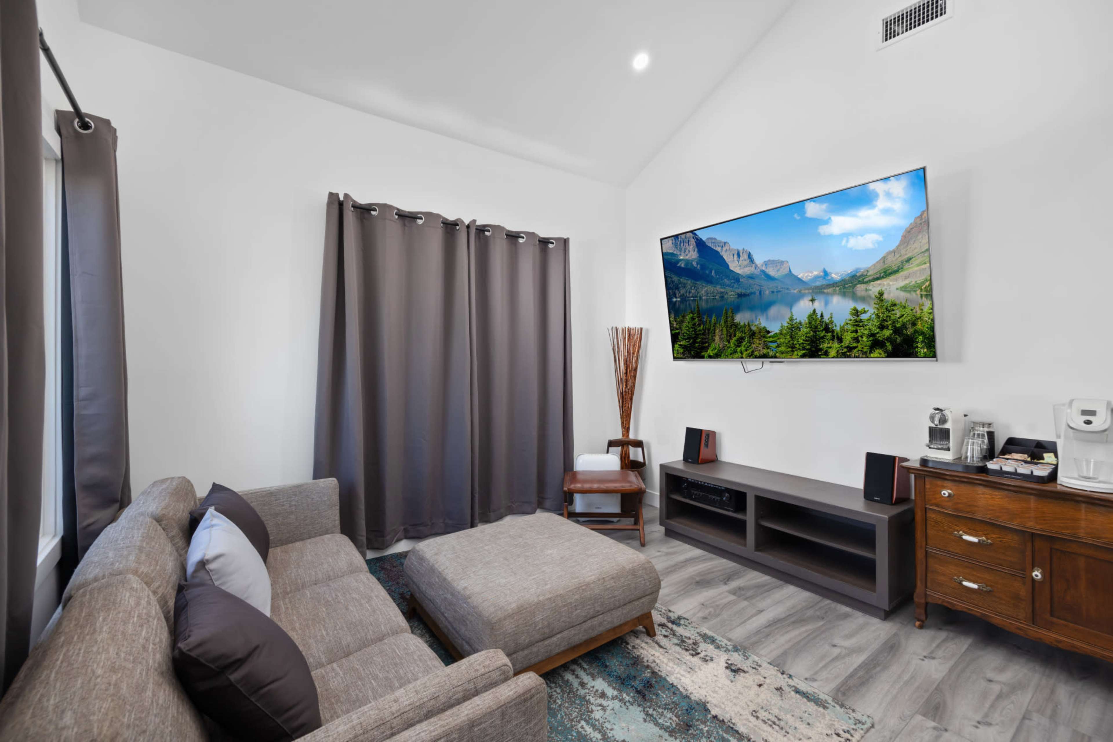 A modern living room with a large television mounted on the wall, a sectional sofa, and a coffee table, all positioned near windows covered with dark curtains.