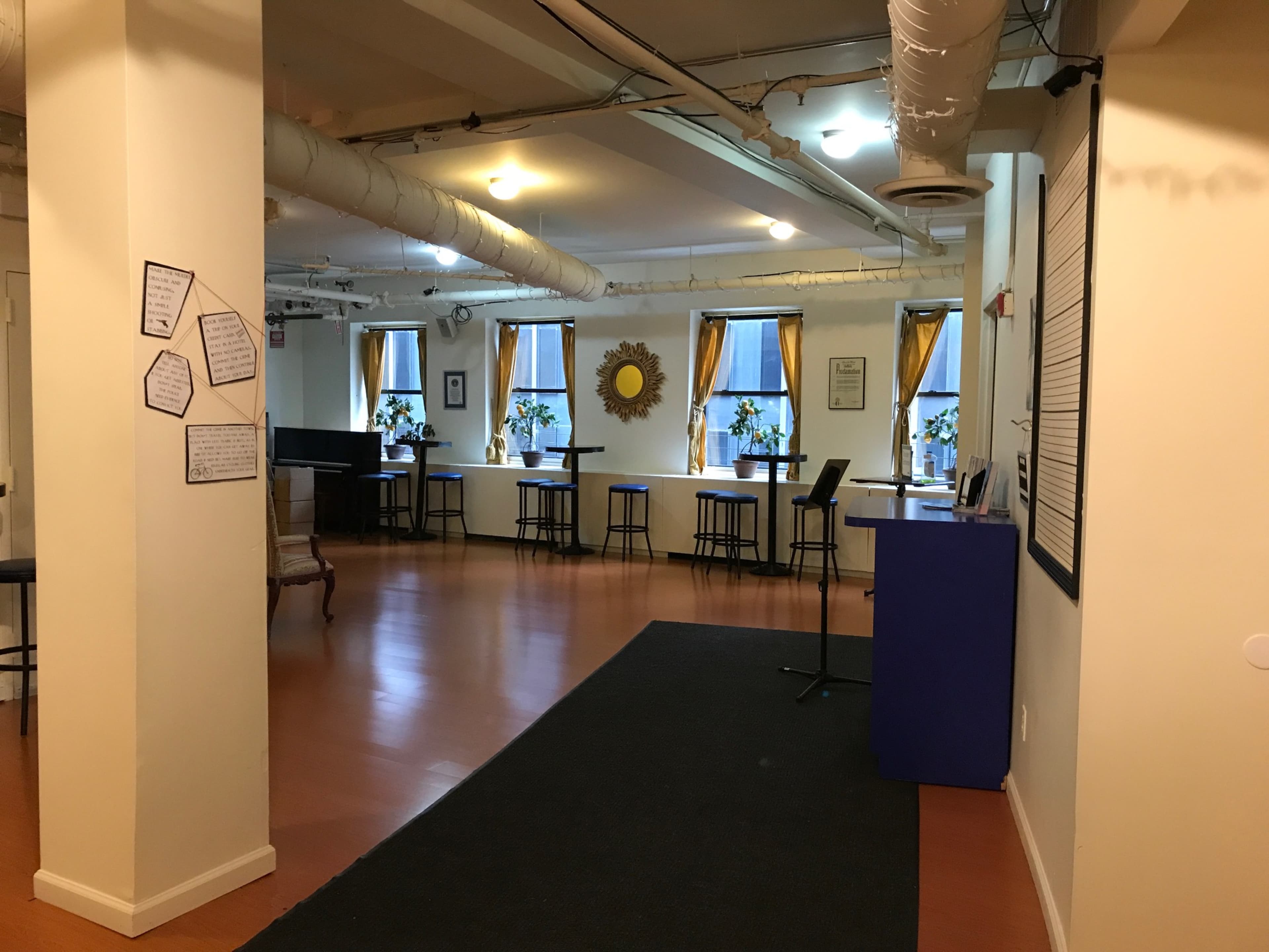 The image shows a spacious room with windows featuring yellow curtains, tables and chairs arranged around the perimeter, and a small bar area on one side.