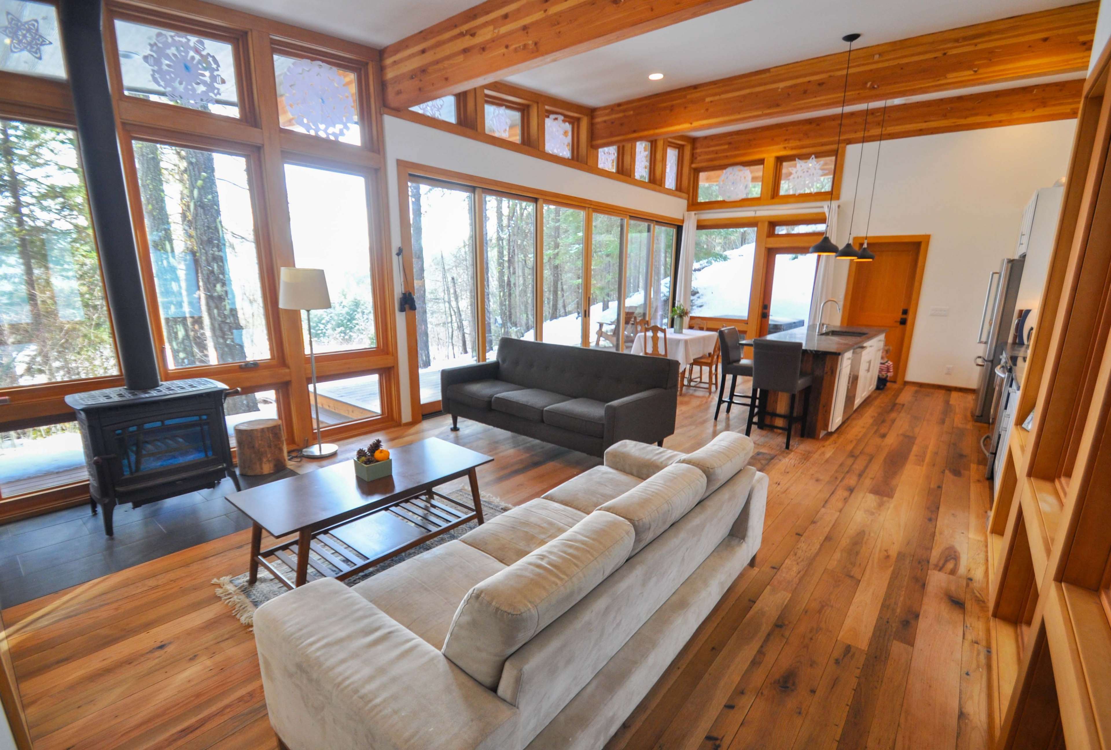 The image shows a living room with large windows, a modern sofa and coffee table, and an open layout leading to a kitchen area, all featuring wooden accents.