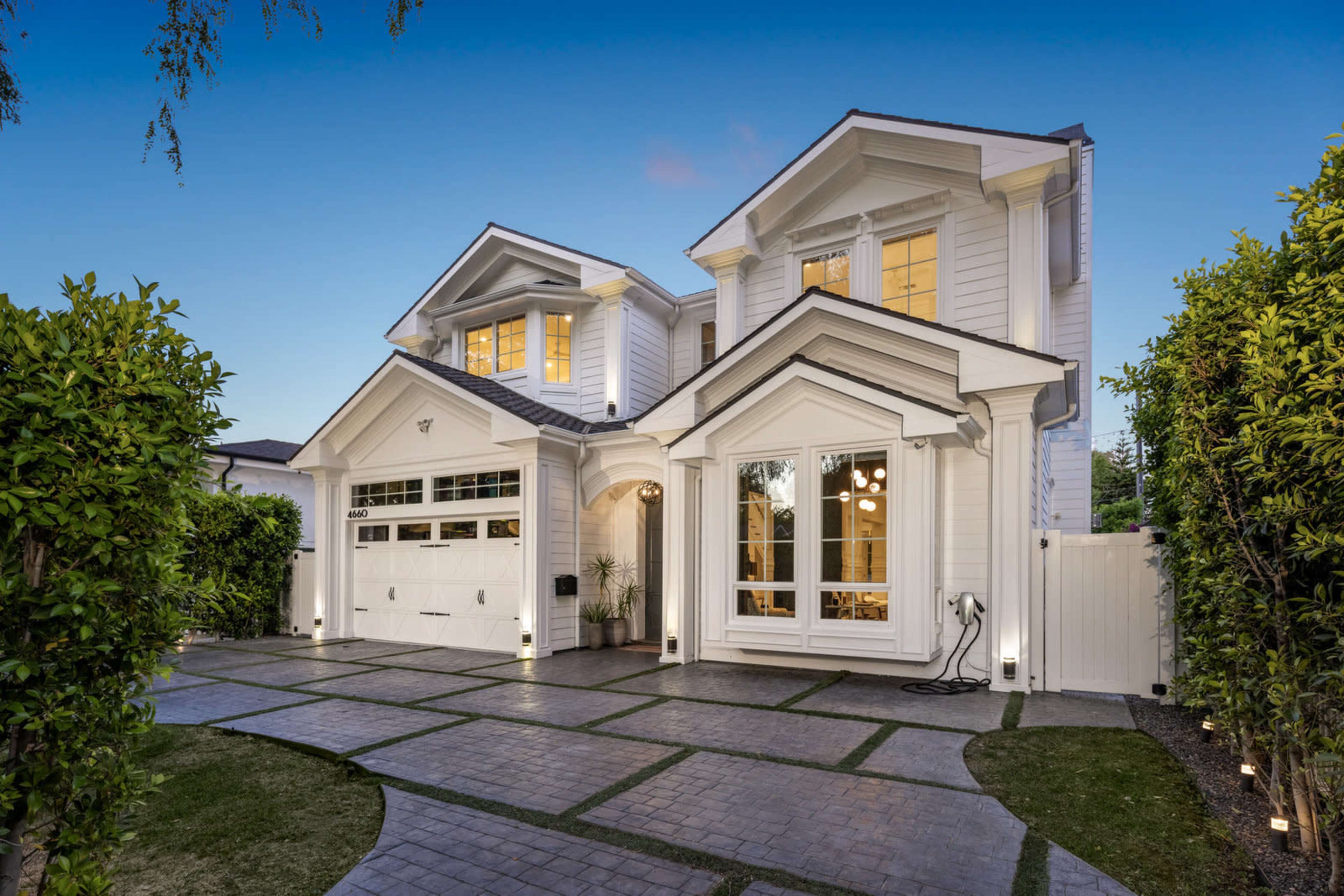 The image shows a modern two-story white house with a paved driveway and landscaped greenery.